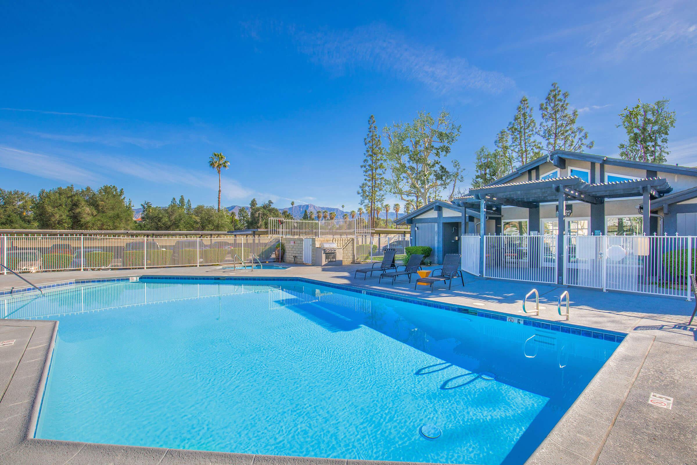 A sparkling blue swimming pool surrounded by a patio area, lounge chairs, and a fenced perimeter. In the background, there are trees and a clear blue sky, creating a relaxing atmosphere. A building with a shaded area is visible near the pool.