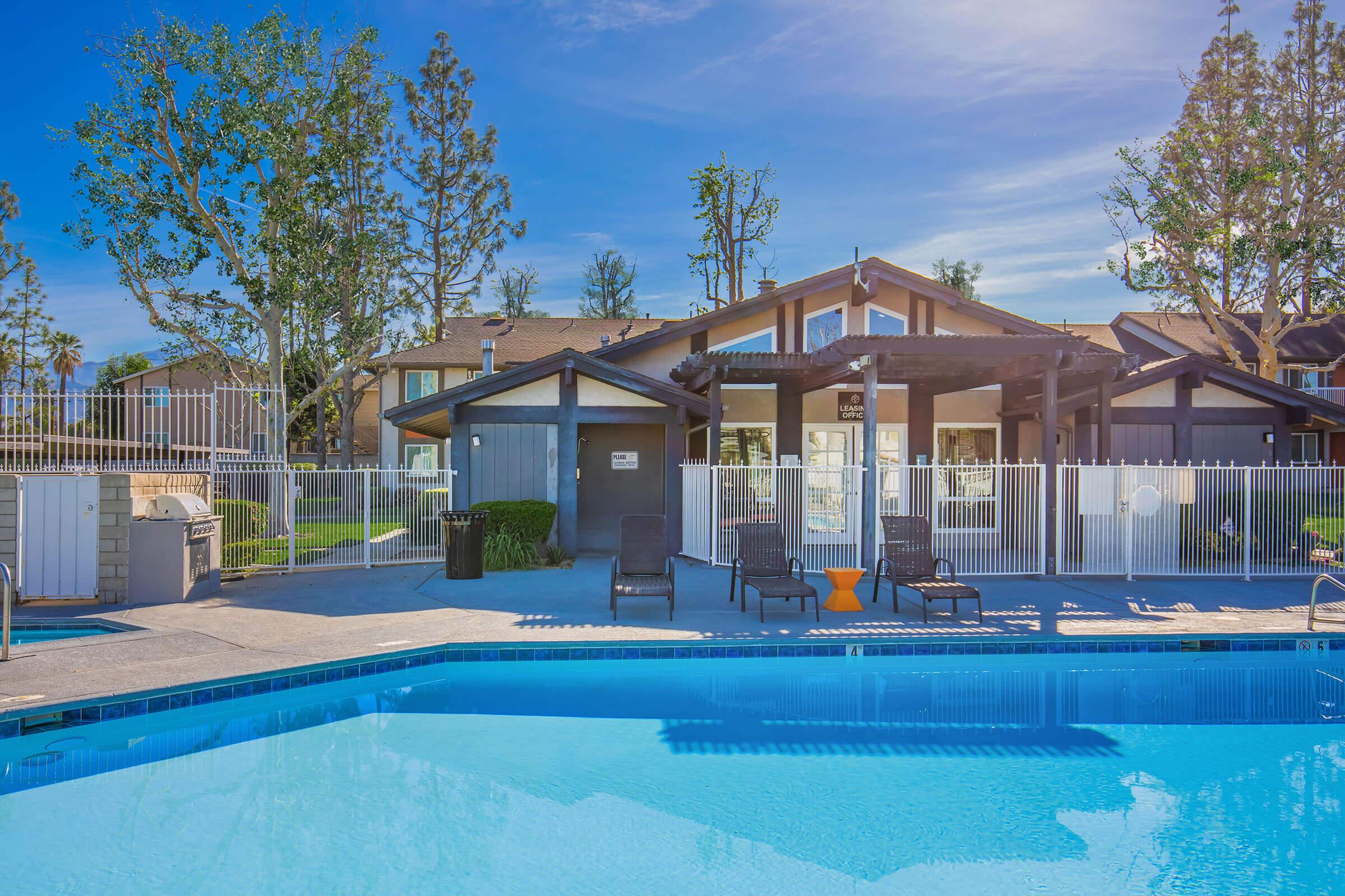 A view of a community swimming pool surrounded by lounge chairs and a fence. In the background, there are modern buildings with a clear blue sky and trees. The pool has a clear, inviting blue water, reflecting the sunlight.