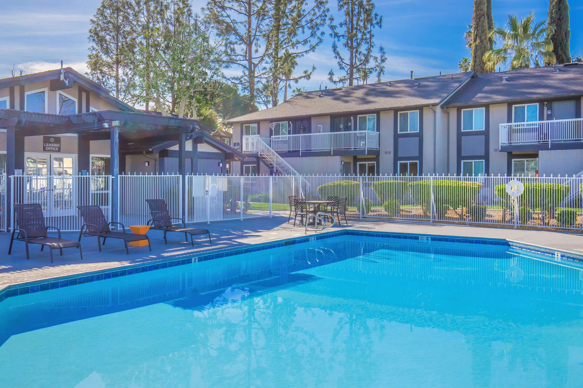 A sunny pool area with clear blue water surrounded by lounge chairs and a fenced patio. In the background, there are several two-story buildings with balconies and lush greenery. The scene conveys a relaxed outdoor atmosphere ideal for leisure or recreation.