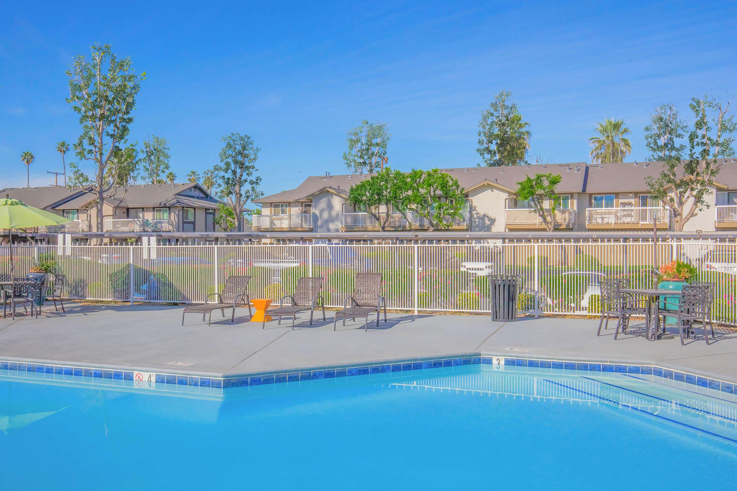 A sunny poolside view featuring a clear blue swimming pool with lounge chairs arranged around it. In the background, there are green trees and residential buildings, creating a relaxing atmosphere. The sky is clear and blue, enhancing the inviting setting.