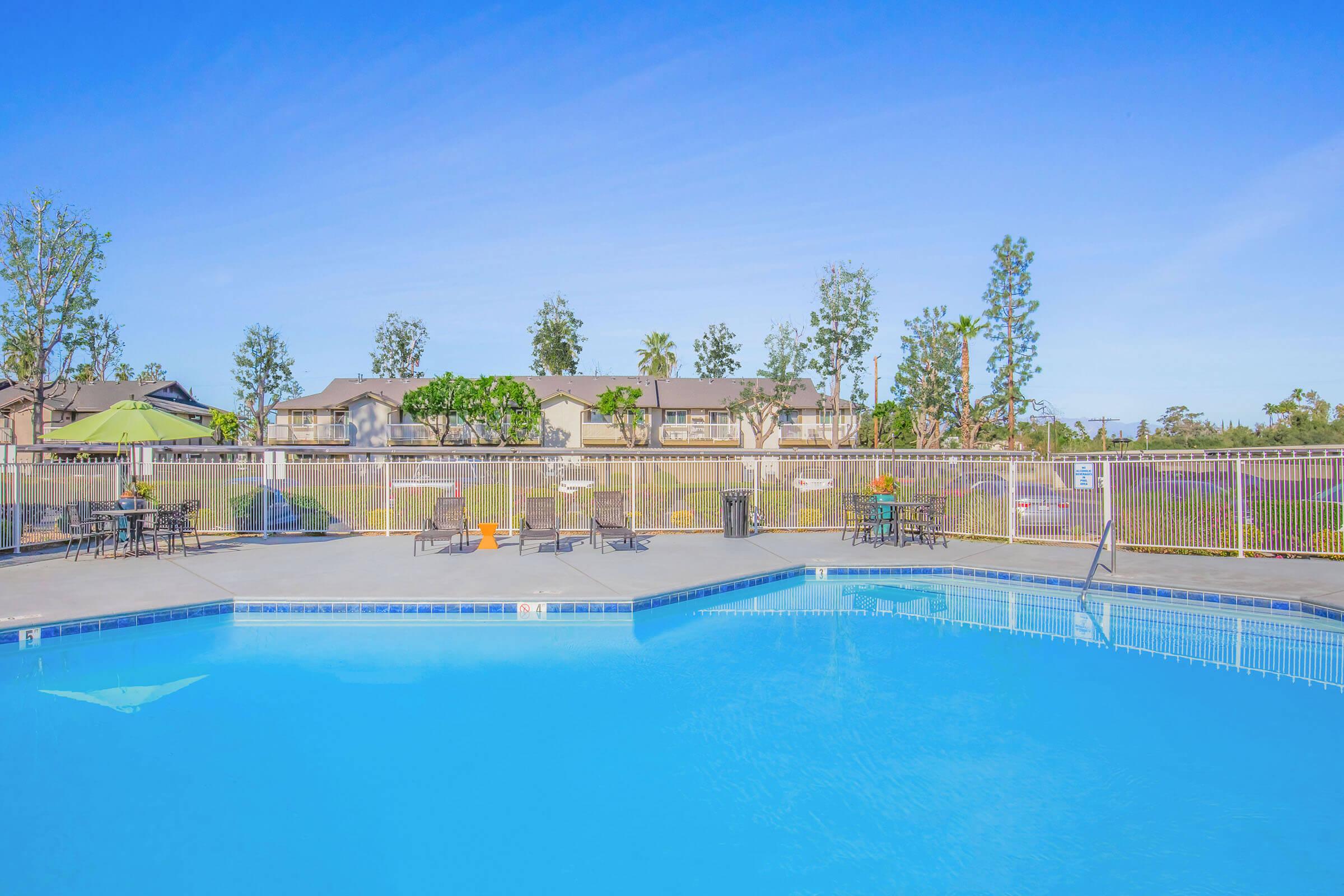 A clear blue swimming pool surrounded by a fence, with several black chairs and tables nearby. Lush green trees and low-rise buildings are visible in the background under a bright blue sky.