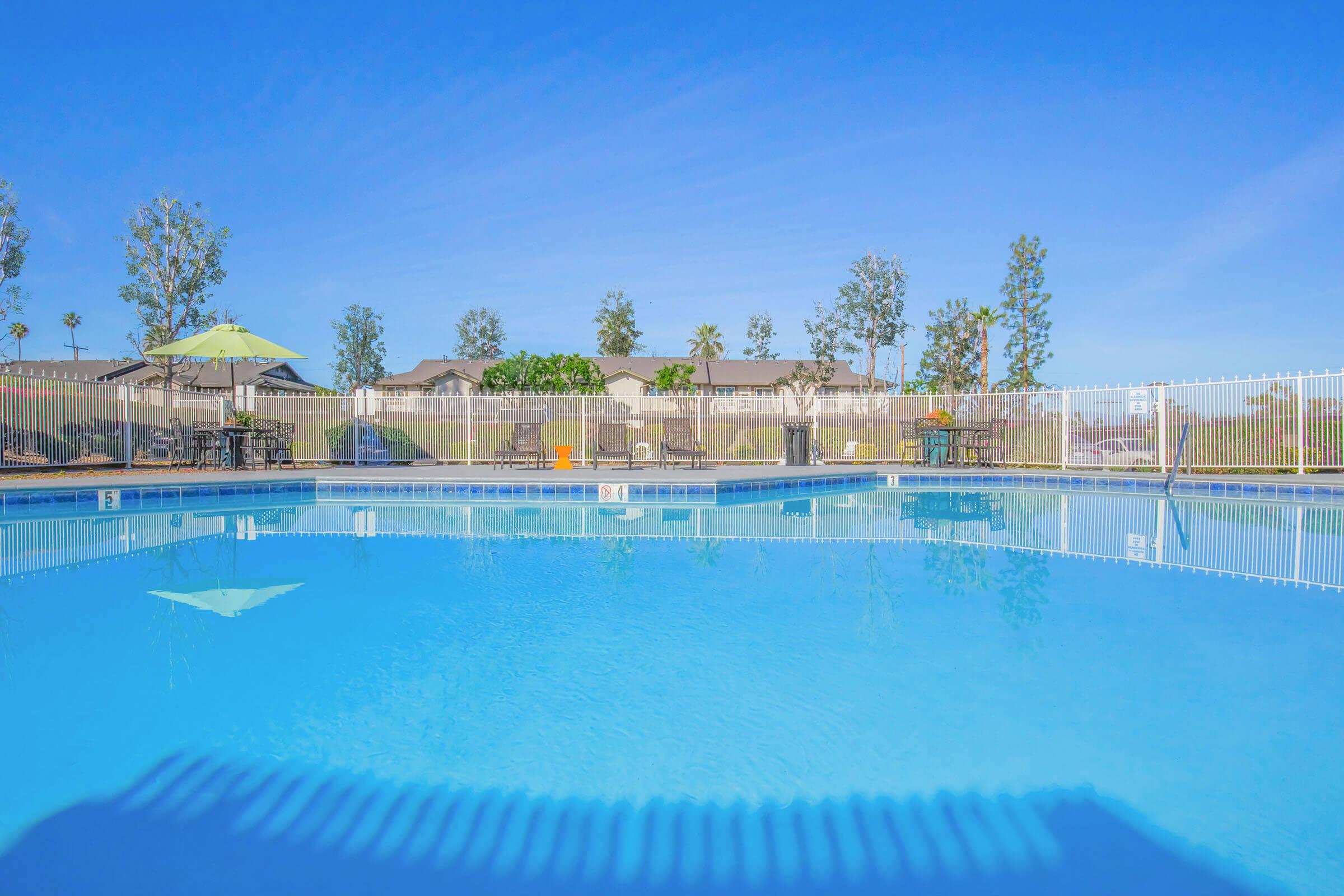 A clear blue swimming pool surrounded by a white fence, with several lounge chairs and tables under umbrellas in the background. Lush trees and residential buildings are visible beyond the pool area, under a bright blue sky.