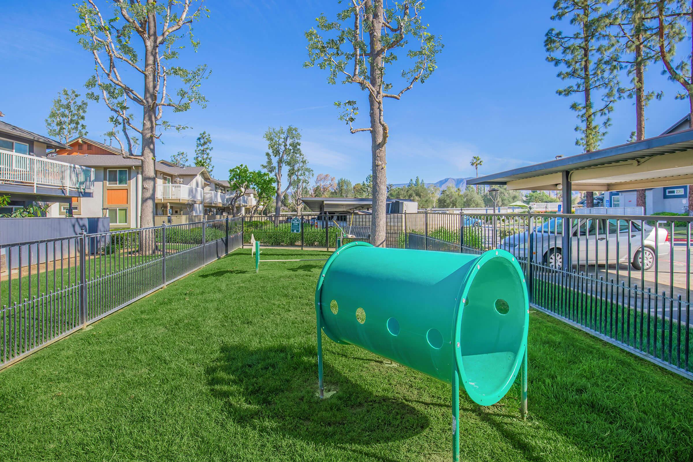 A sunny park area featuring a vibrant green play tunnel on grass, surrounded by trees and modern residential buildings. The space is fenced, and there are additional play equipment elements visible, creating an inviting atmosphere for outdoor activities.