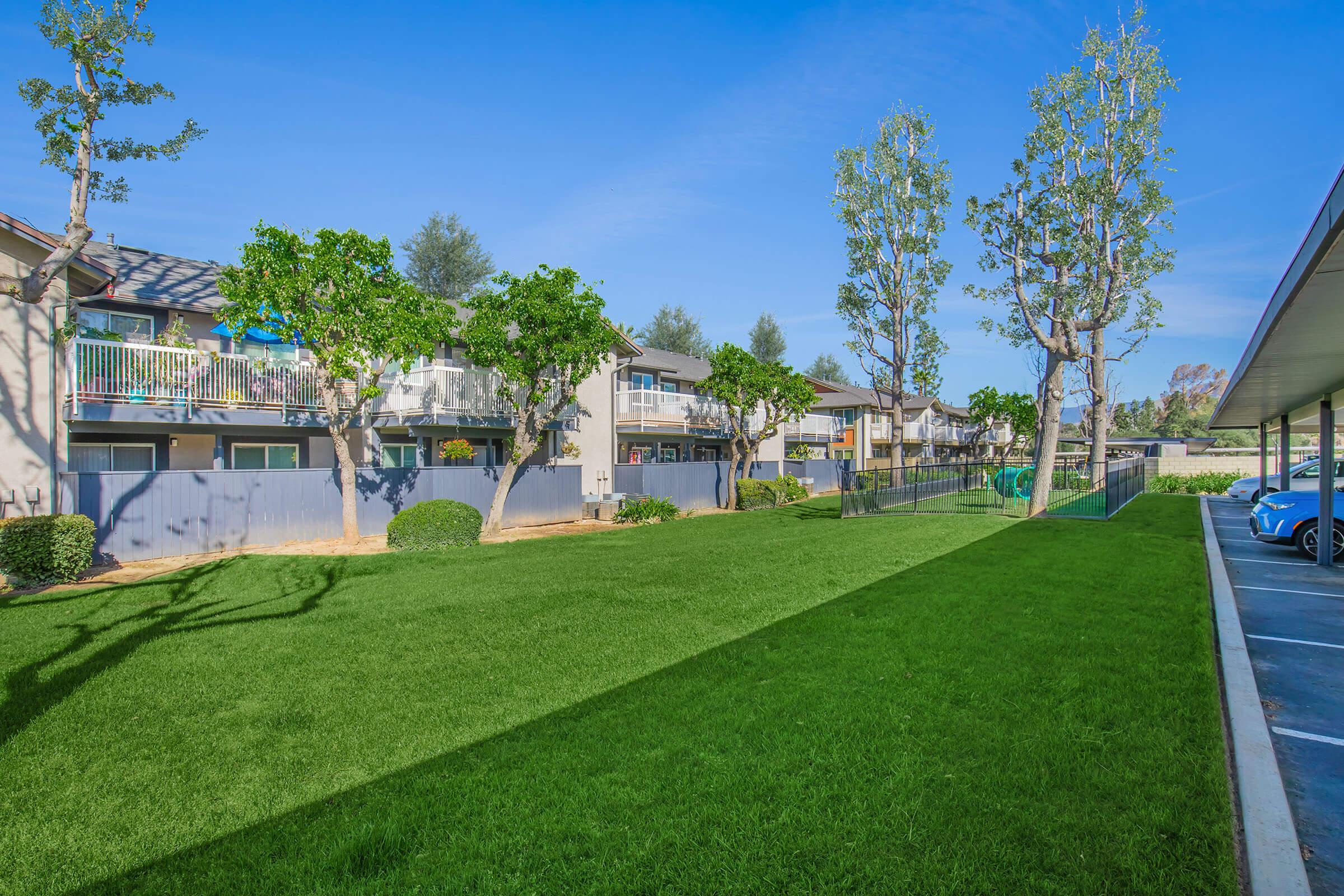 Lush green lawn in front of apartment buildings, framed by trees. The buildings have balconies and are surrounded by landscaped grounds. A clear blue sky is visible above, creating a bright and inviting atmosphere.