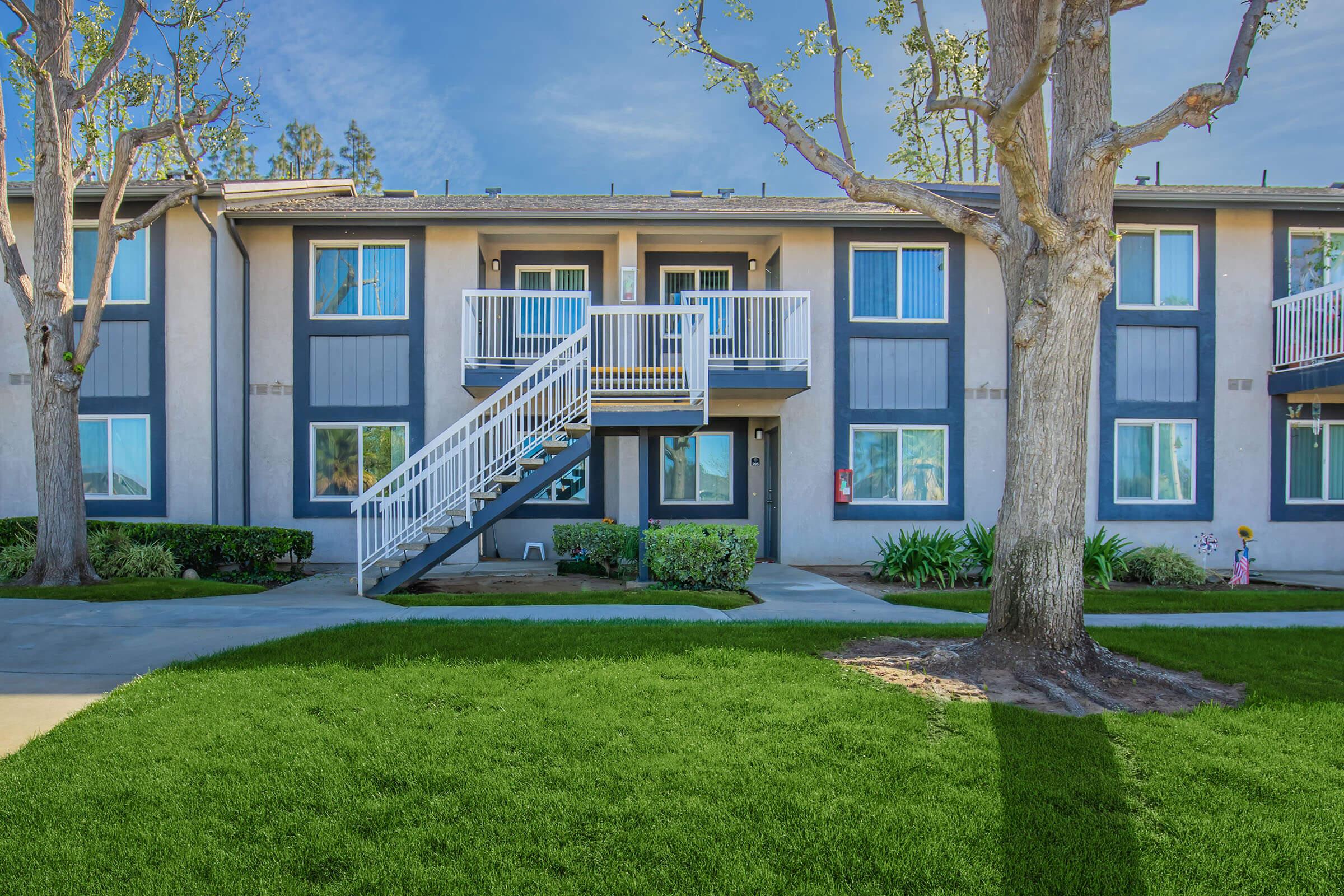 A view of an apartment building with a white staircase leading to the second floor. The building features blue windows, green grass, and a tree in the foreground. The surroundings are well-maintained, with shrubs and a clear blue sky in the background.