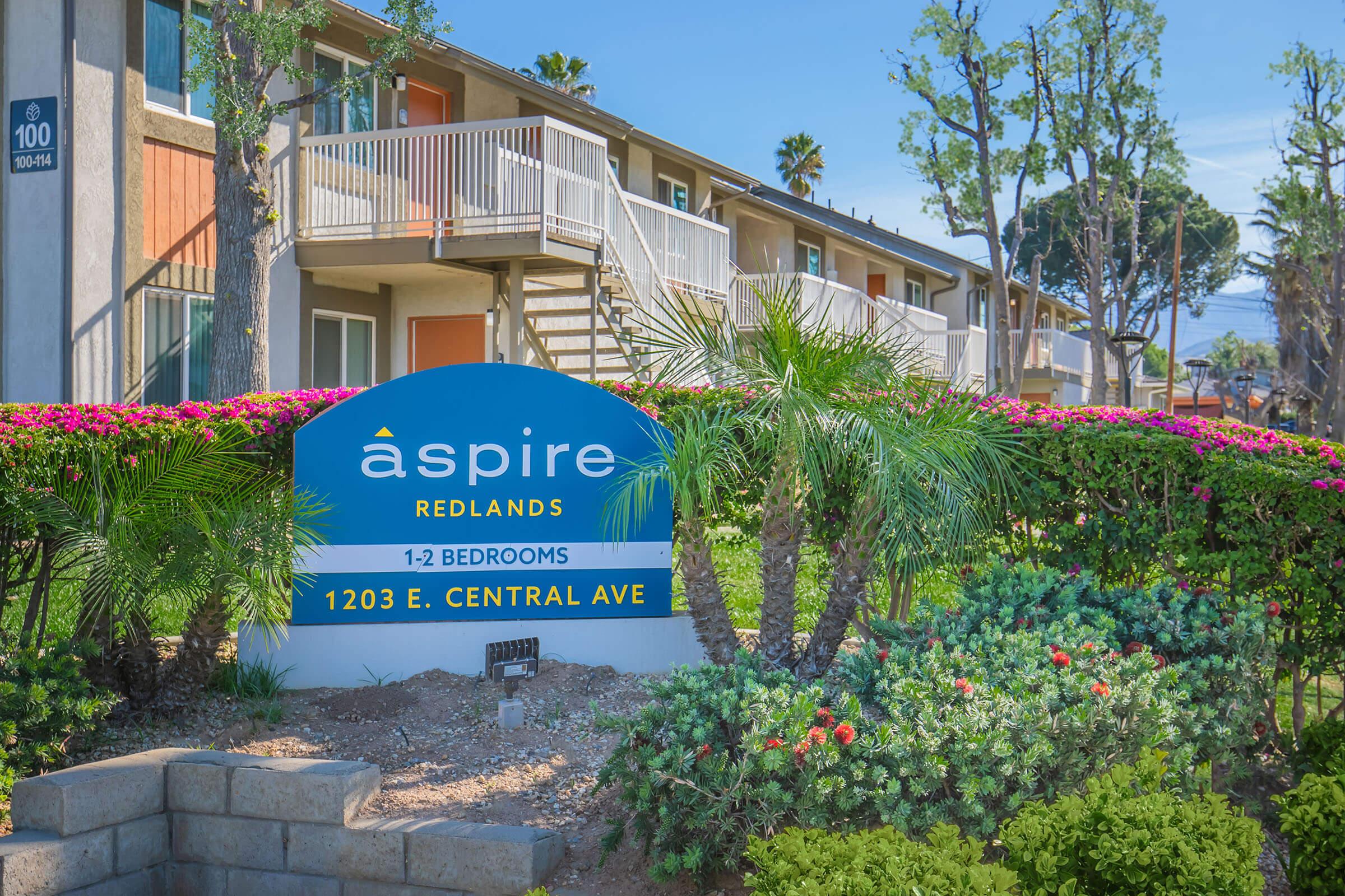 Sign for Aspire Redlands apartments, featuring "1-2 Bedrooms" and the address "1203 E. Central Ave." Lush green landscaping and colorful flowers surround the sign, with residential buildings in the background under a clear blue sky.