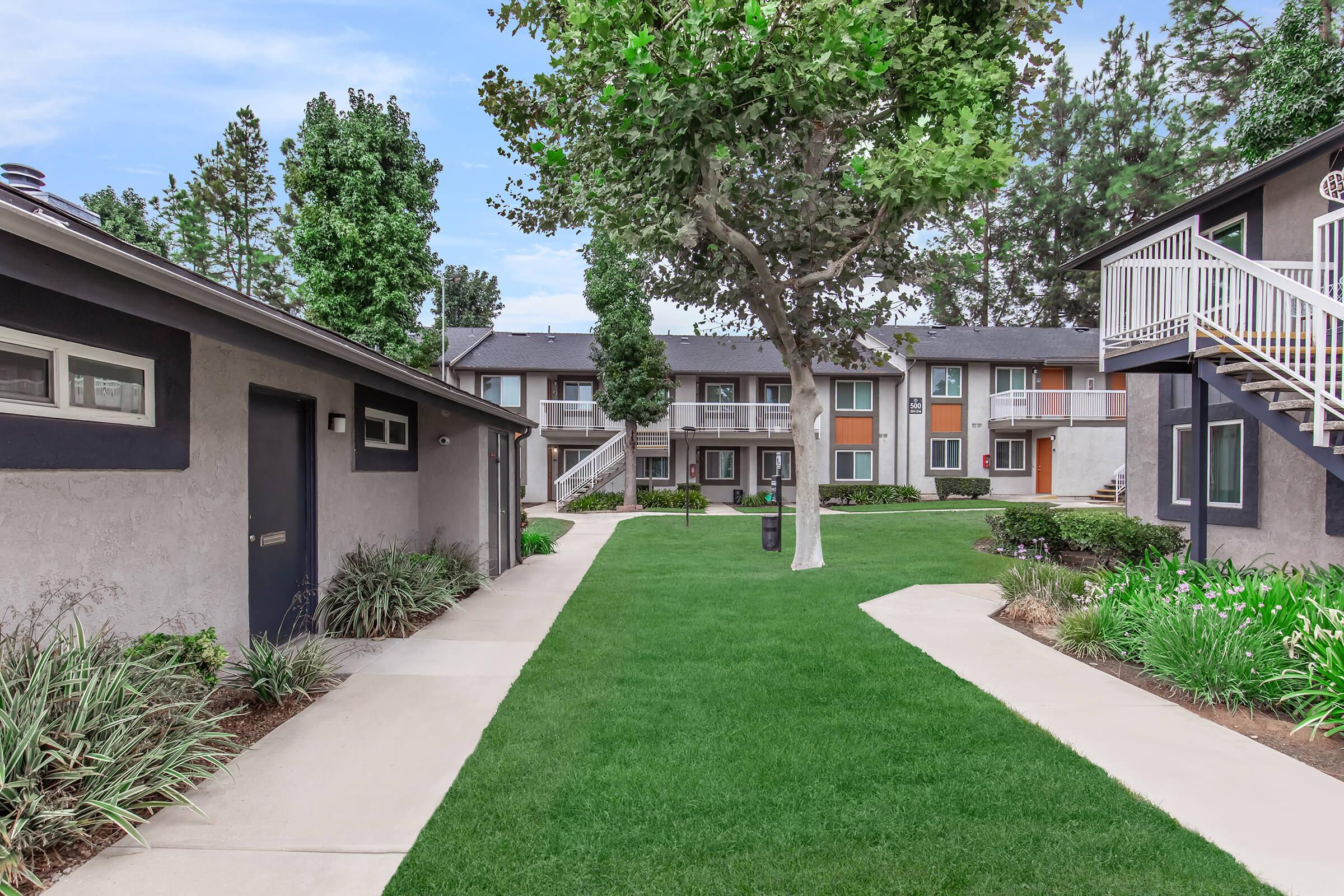 A green lawn surrounded by buildings, featuring pathways, trees, and landscaped areas. There are two buildings visible with balconies, and a well-maintained outdoor space providing a peaceful residential environment.