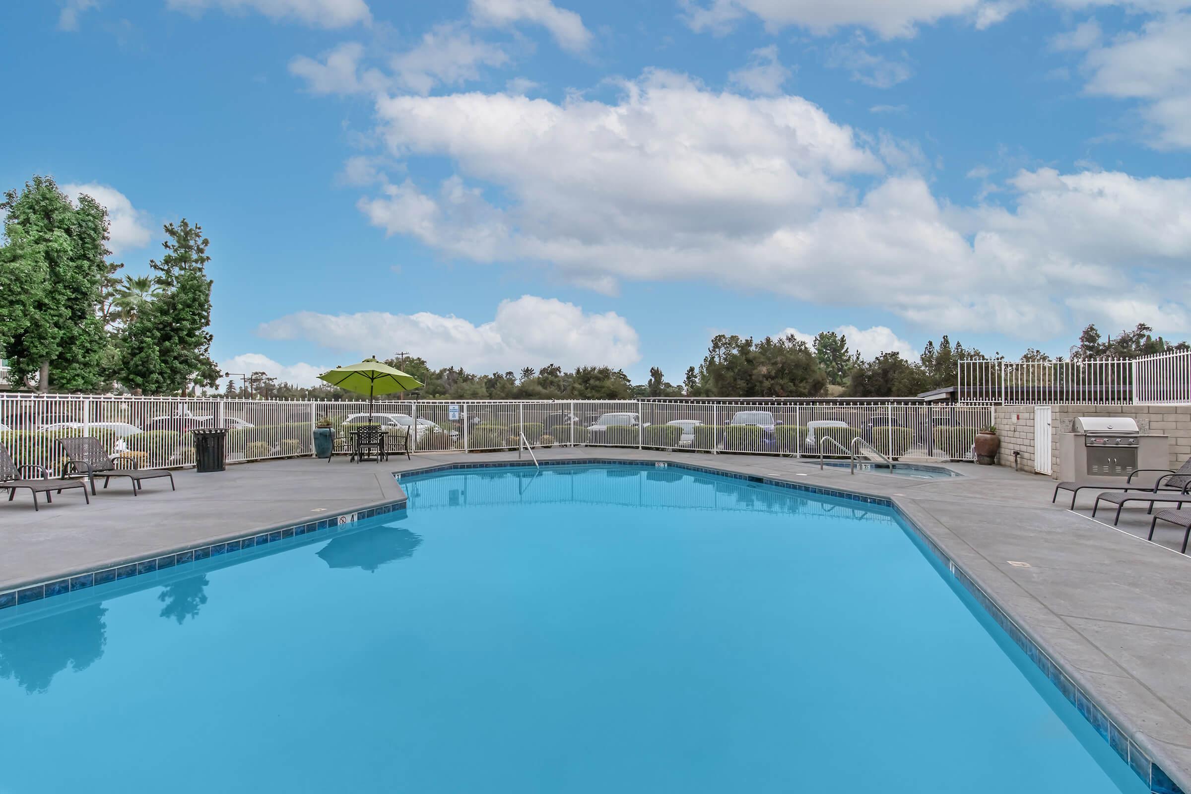 A clear blue swimming pool surrounded by lounge chairs and tables. In the background, there are trees and a cloudy sky. A green umbrella is positioned near the pool's edge, creating a relaxing outdoor environment.