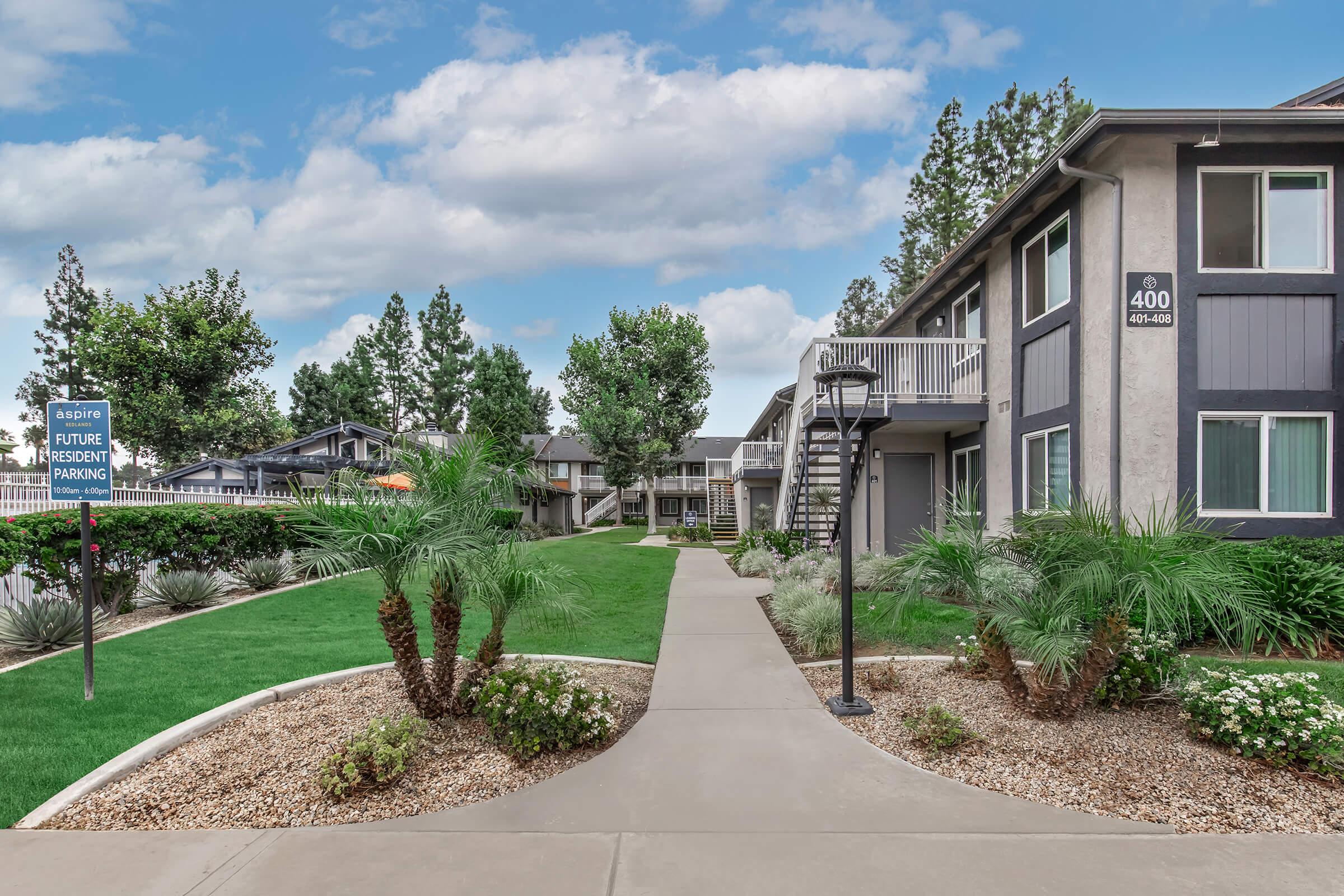 A landscaped apartment complex featuring walking paths, manicured lawns, and palm trees. Multiple buildings are visible, with a sign for future resident parking. The scene is set against a backdrop of blue skies and scattered clouds.