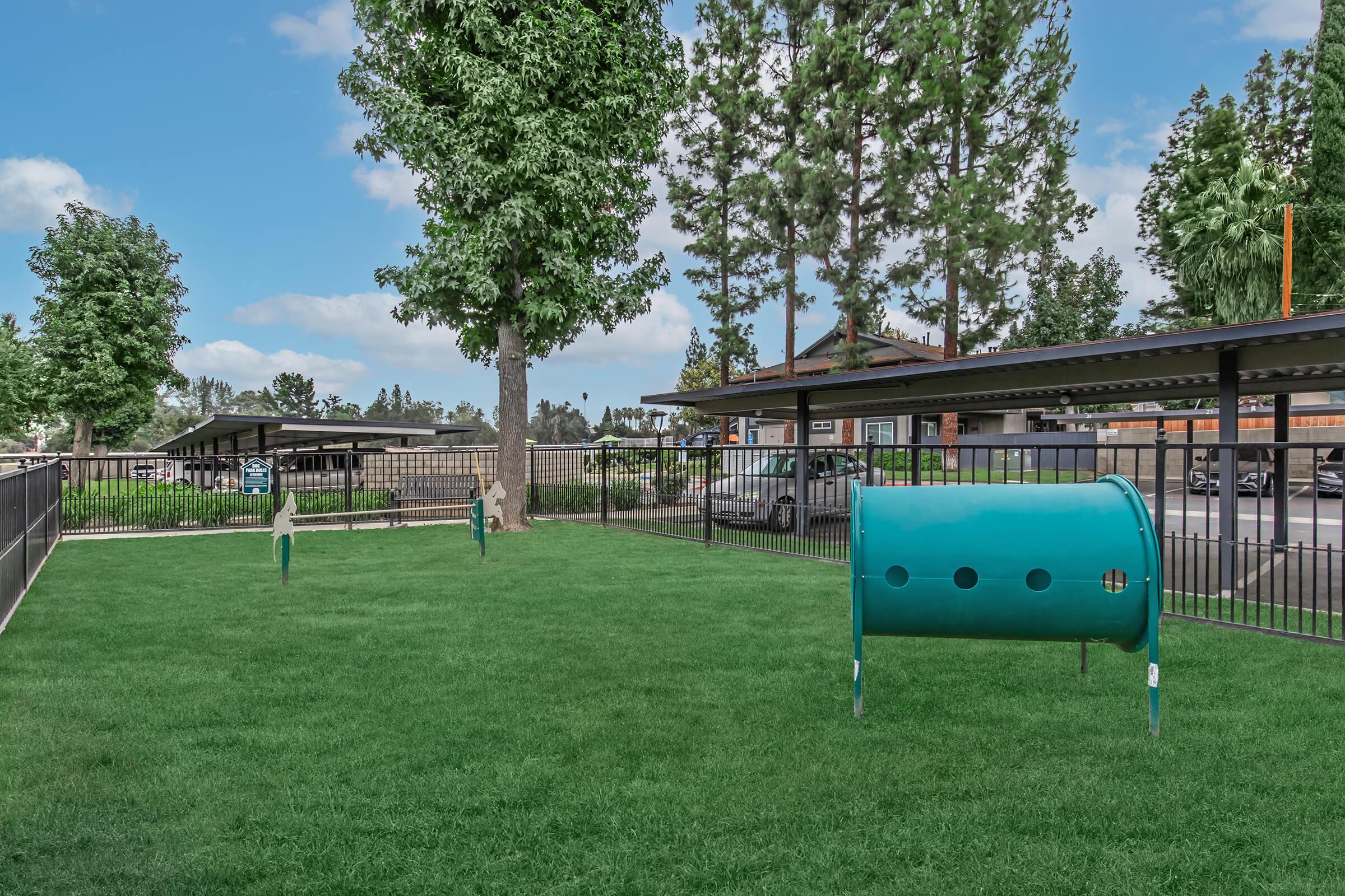 A green grassy area enclosed by a black fence, featuring two playful equipment structures and a large blue tunnel. In the background, there are trees and a parking area with cars under shaded structures. The scene is bright and inviting, ideal for outdoor activities.
