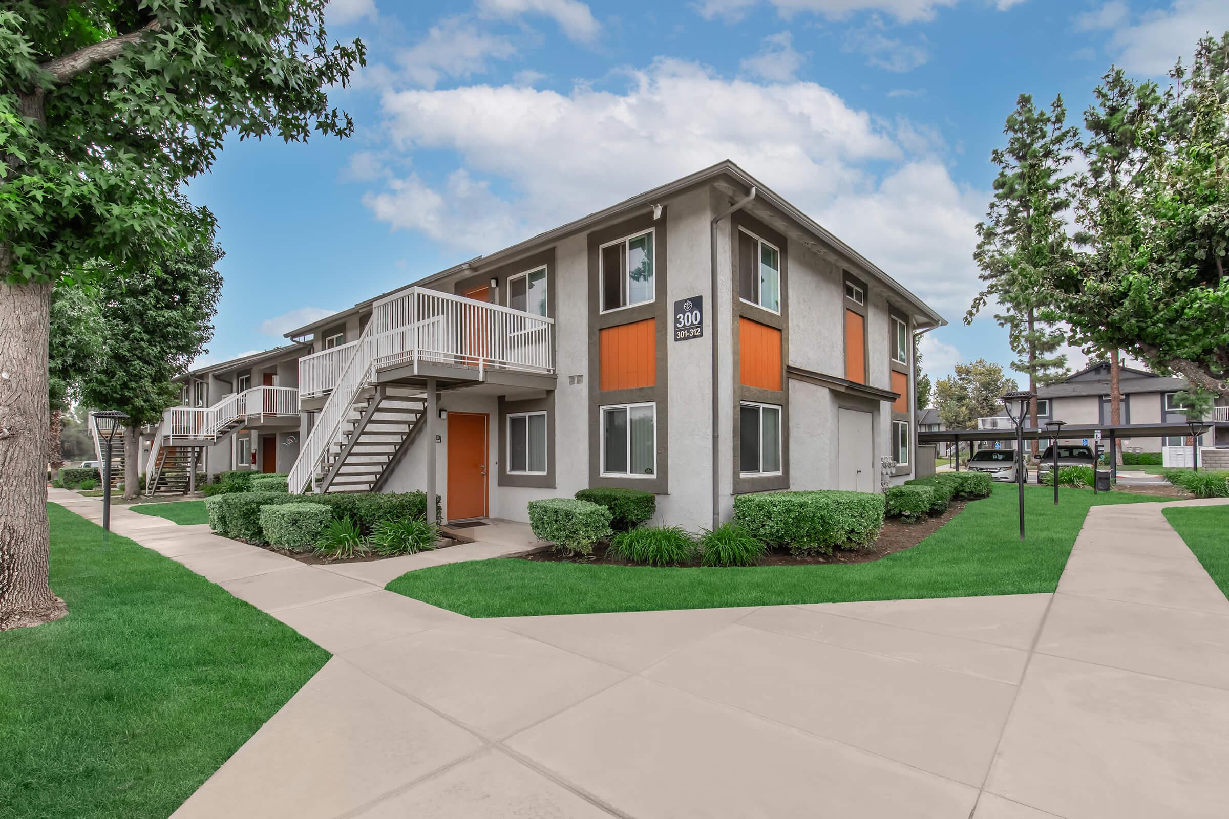 A two-story residential apartment building with a light grey exterior and orange accents. The building features a staircase on one side, surrounded by neatly landscaped green lawns and shrubs. A paved pathway leads to the entrance, with a bright blue sky in the background.