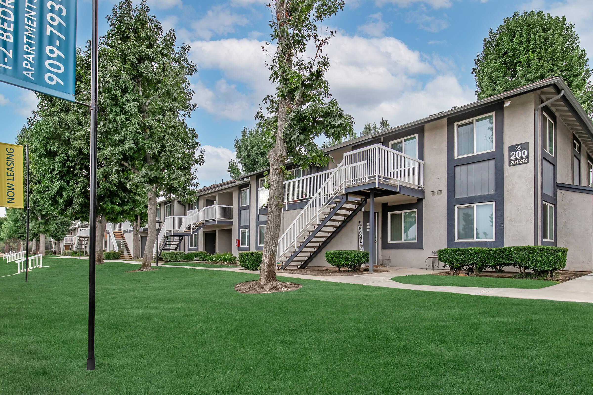 Two-story apartment buildings with gray exteriors, white railings, and green landscaping. A "Now Leasing" sign is visible in front of the buildings, along with well-maintained grass and trees lining the walkway. The sky is partly cloudy, creating a bright atmosphere.