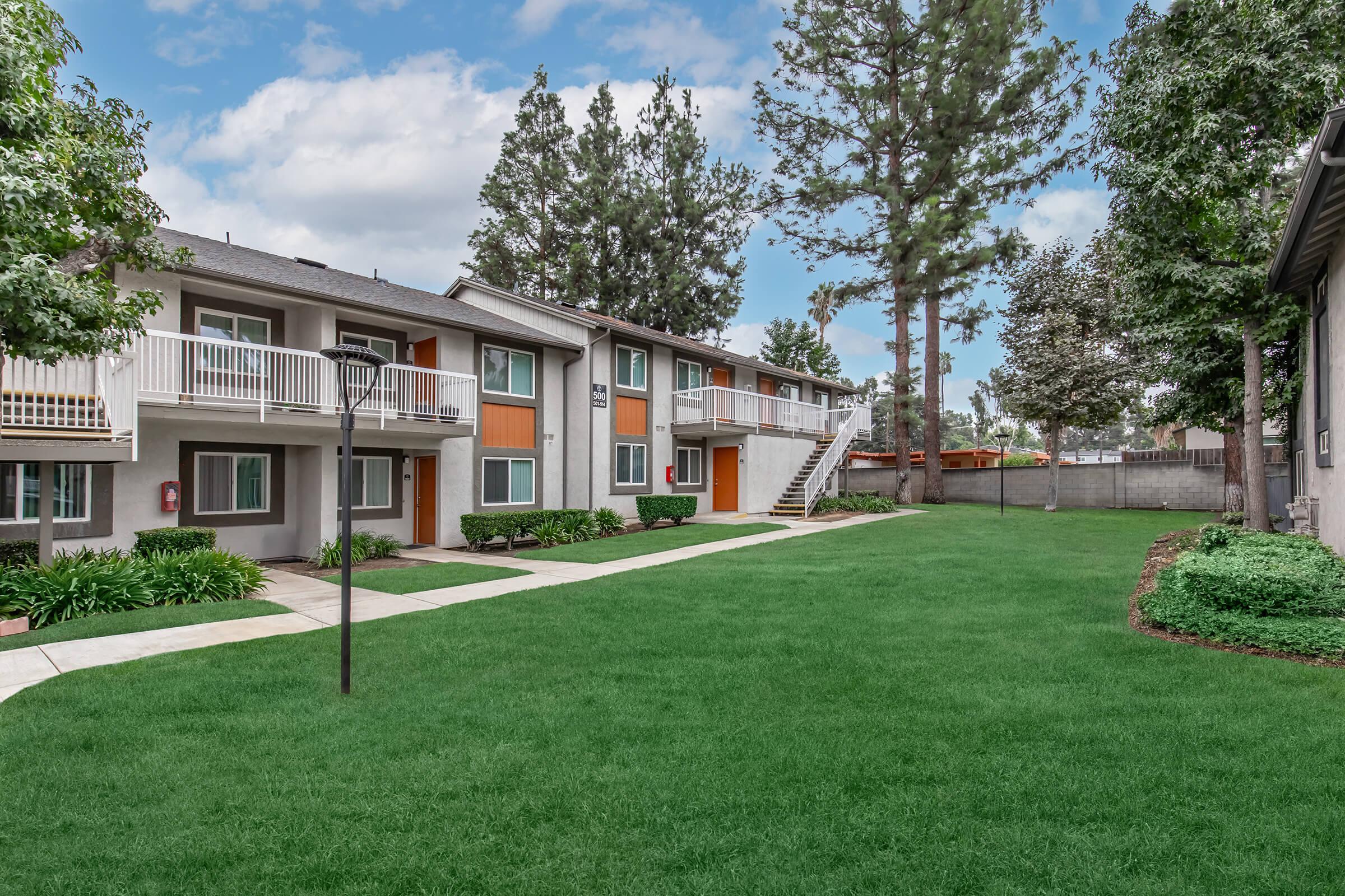 A well-maintained apartment complex with two-story buildings surrounded by lush green grass and trees. The pathway leads to multiple units, featuring balconies and colorful accents. A clothesline is visible in the yard, adding a homey touch to the outdoor area.