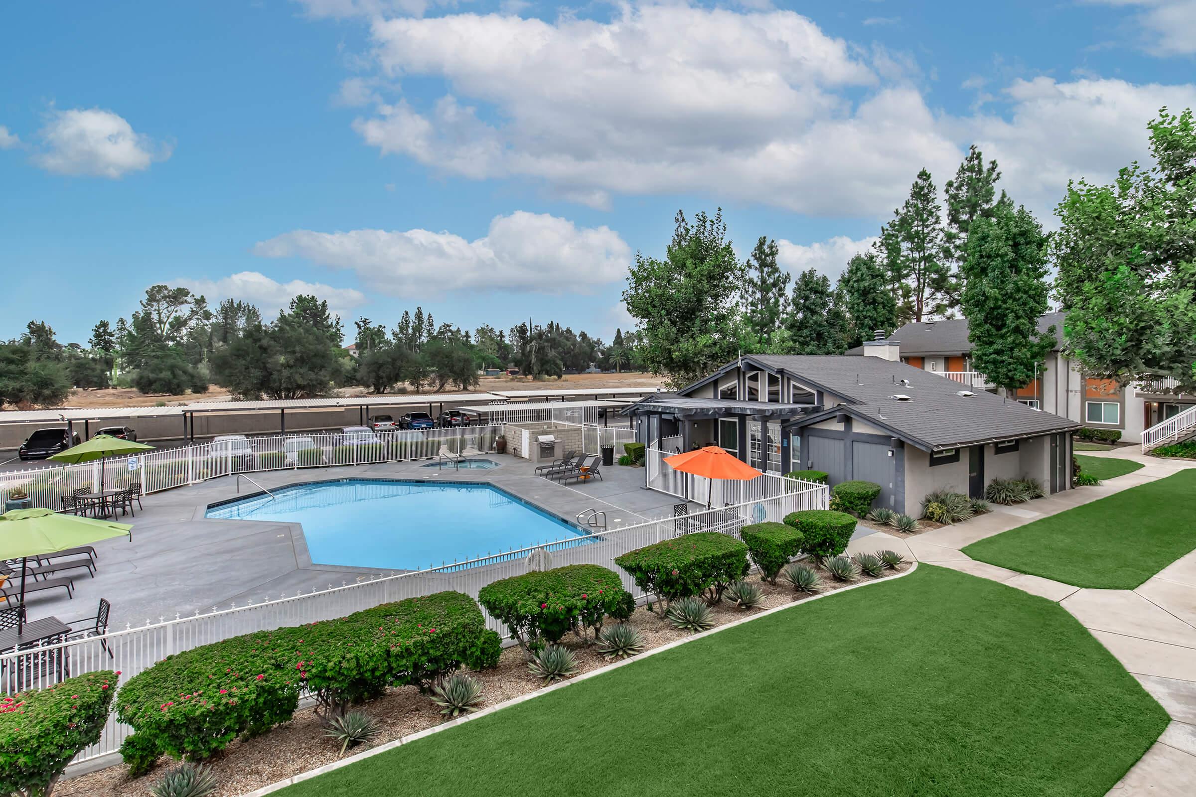 A well-maintained outdoor pool area surrounded by lush greenery and neatly trimmed shrubs. There are lounge chairs and umbrellas near the pool, with a building nearby that provides shelter and amenities. The setting is serene with a clear blue sky and trees in the background.