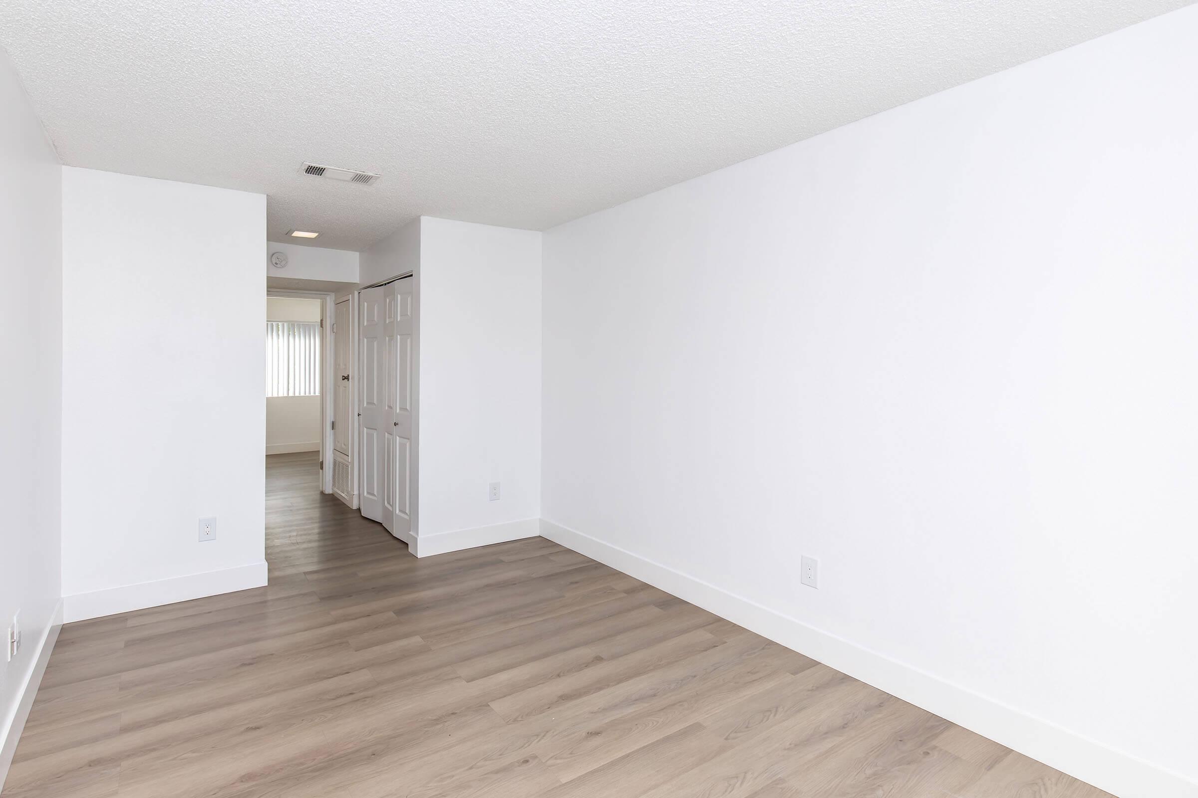 A clean, modern interior of an empty room featuring light-colored walls, wood-like flooring, and a doorway with white double doors leading to another room. Natural light enters through a window with vertical blinds. The space is minimalist and ready for furnishing.