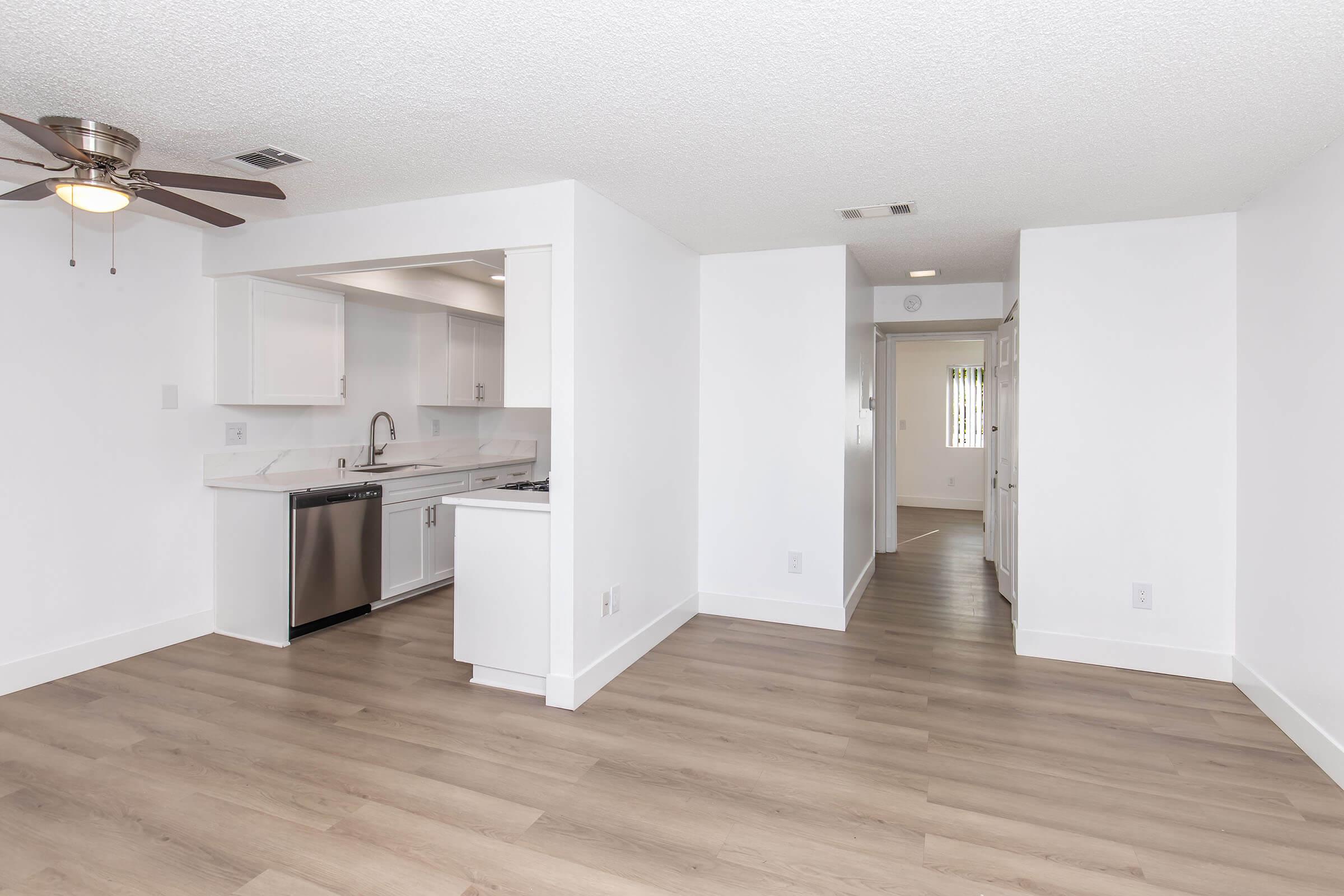 Modern open-concept living space featuring a kitchen with stainless steel appliances, white cabinetry, and a ceiling fan. Light wood flooring extends throughout the area, creating a bright and airy atmosphere. A hallway leads to additional rooms, with natural light illuminating the space.
