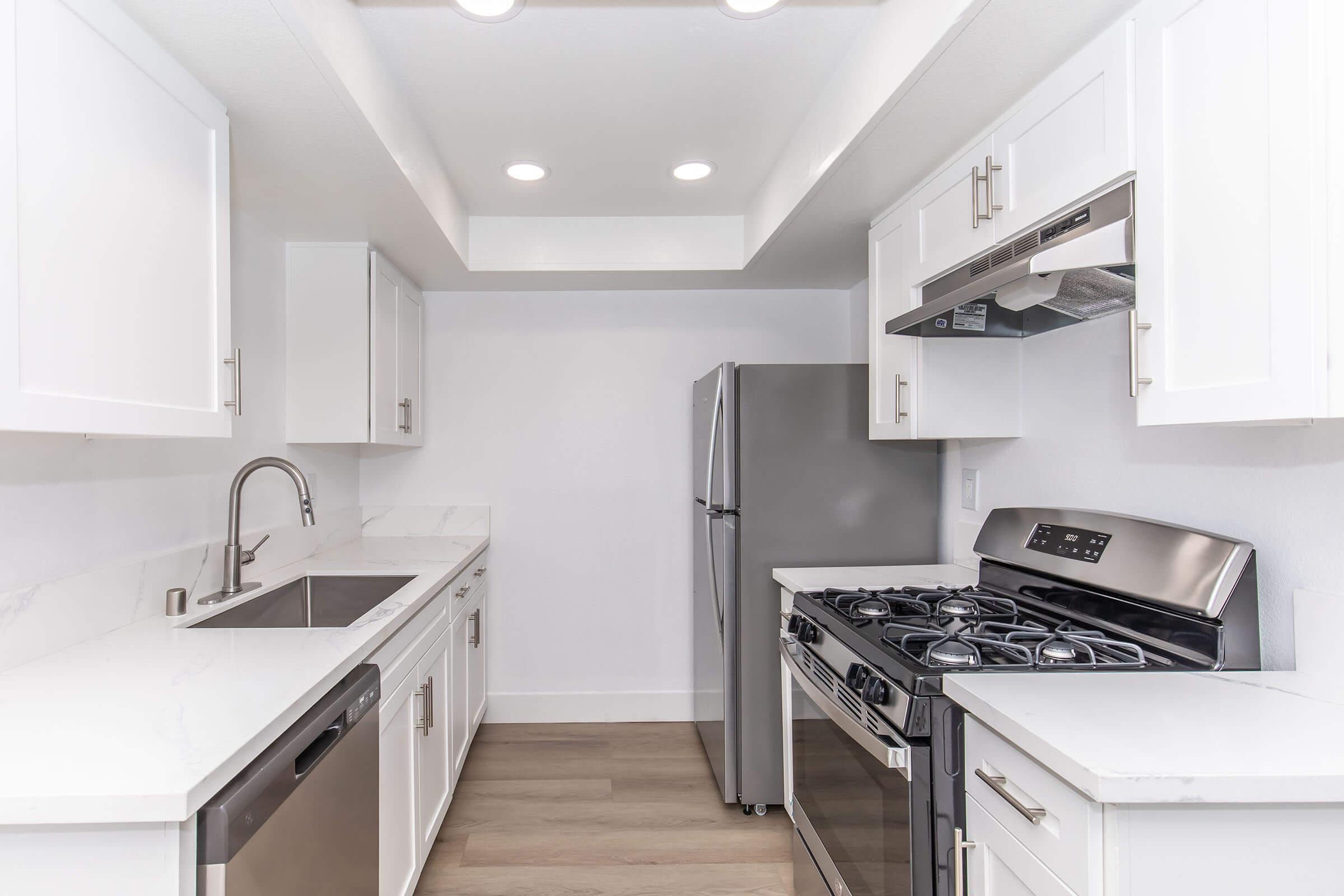 Modern kitchen featuring white cabinets, a stainless steel refrigerator, a gas stove with an oven, and a sleek sink. The countertops are made of white marble, and the space is well-lit with recessed lighting. The kitchen has a clean, contemporary design with a muted color palette.
