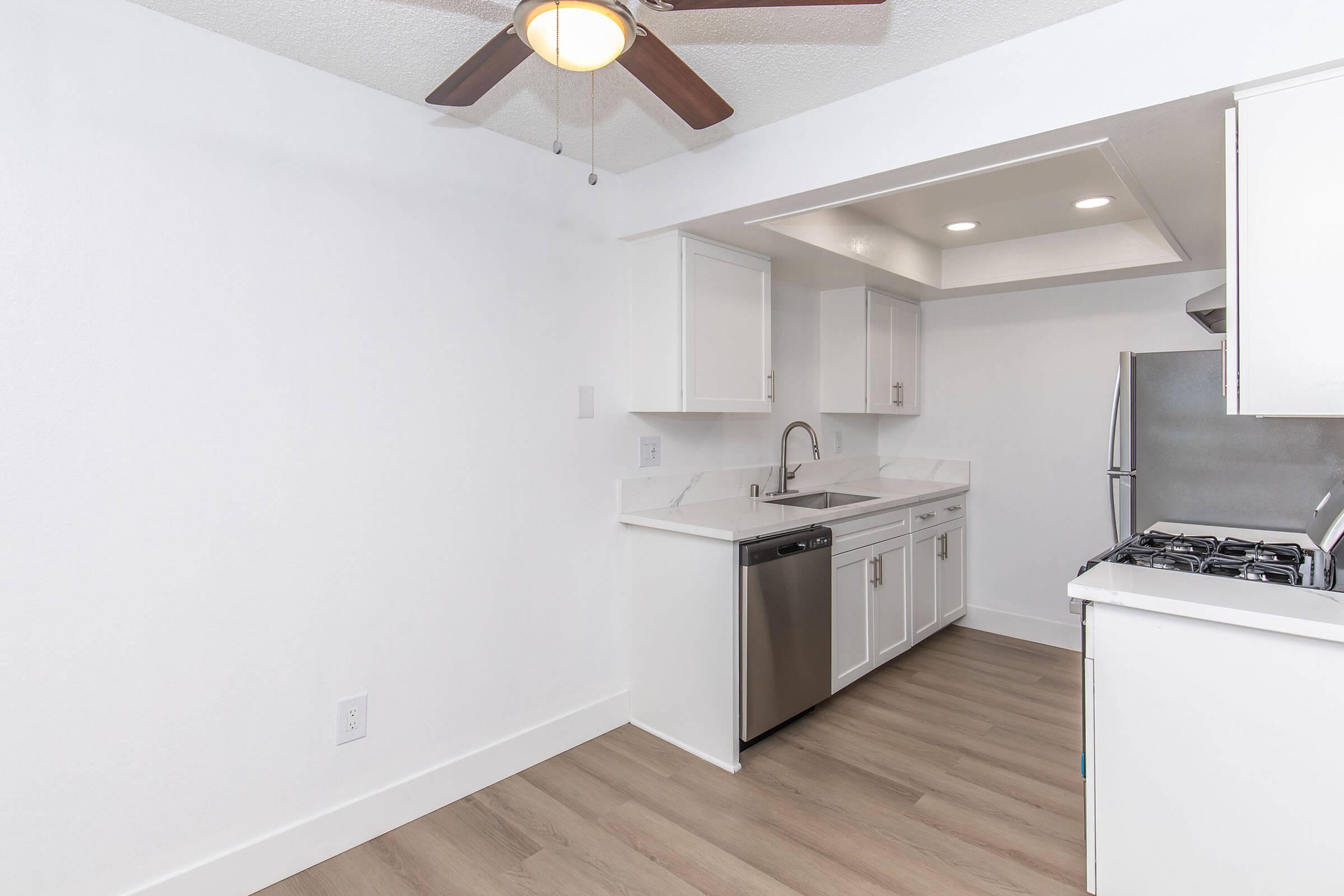 Modern kitchen with white cabinetry, stainless steel appliances, a dishwasher, and a gas stove. The space features a ceiling fan and light fixtures, along with light wood flooring. The walls are painted white, creating a clean and bright atmosphere.