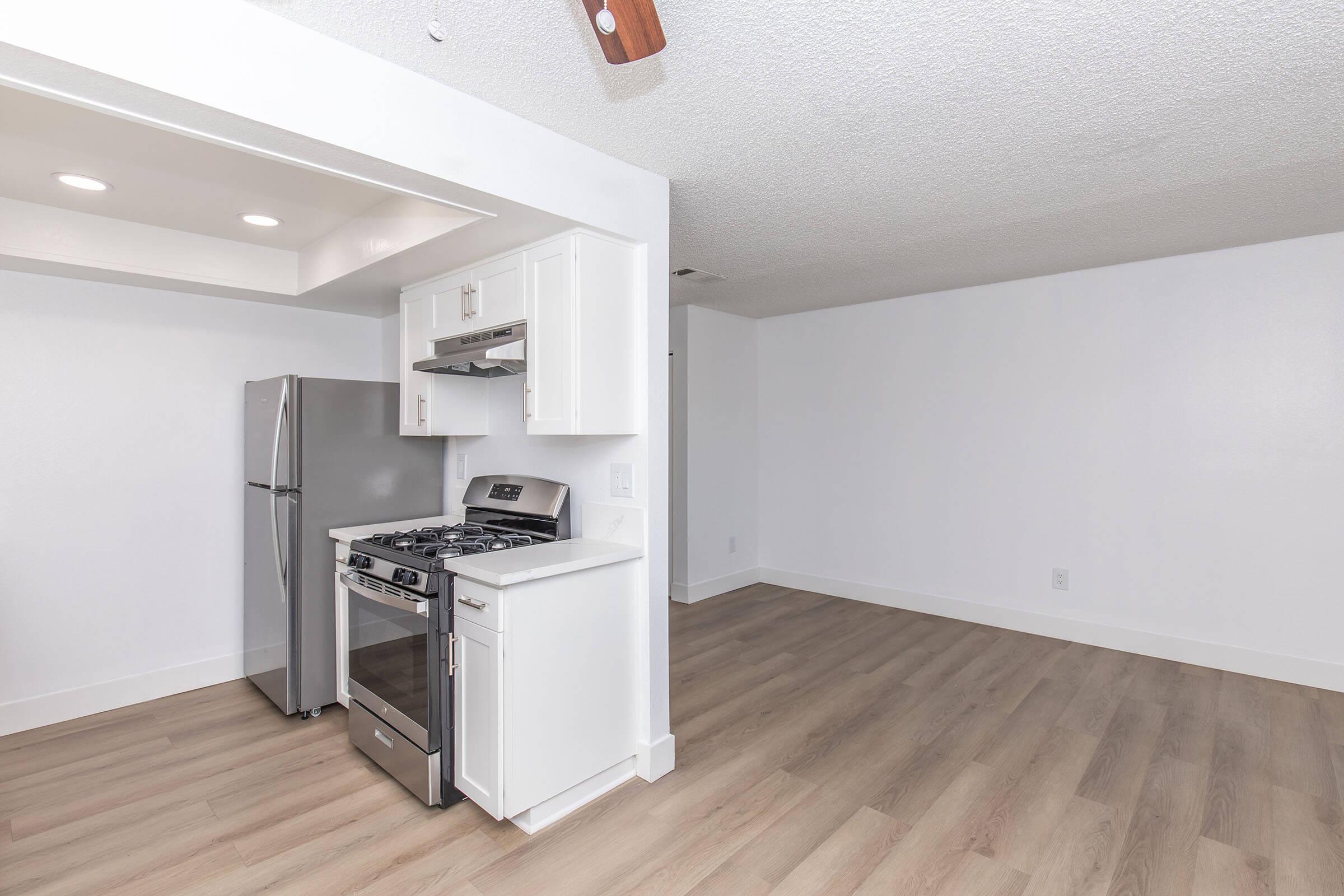 A modern kitchen with stainless steel appliances, including a refrigerator and gas stove, and white cabinetry. The open floor plan features light wood flooring and a bright, neutral color scheme, leading into a spacious living area.