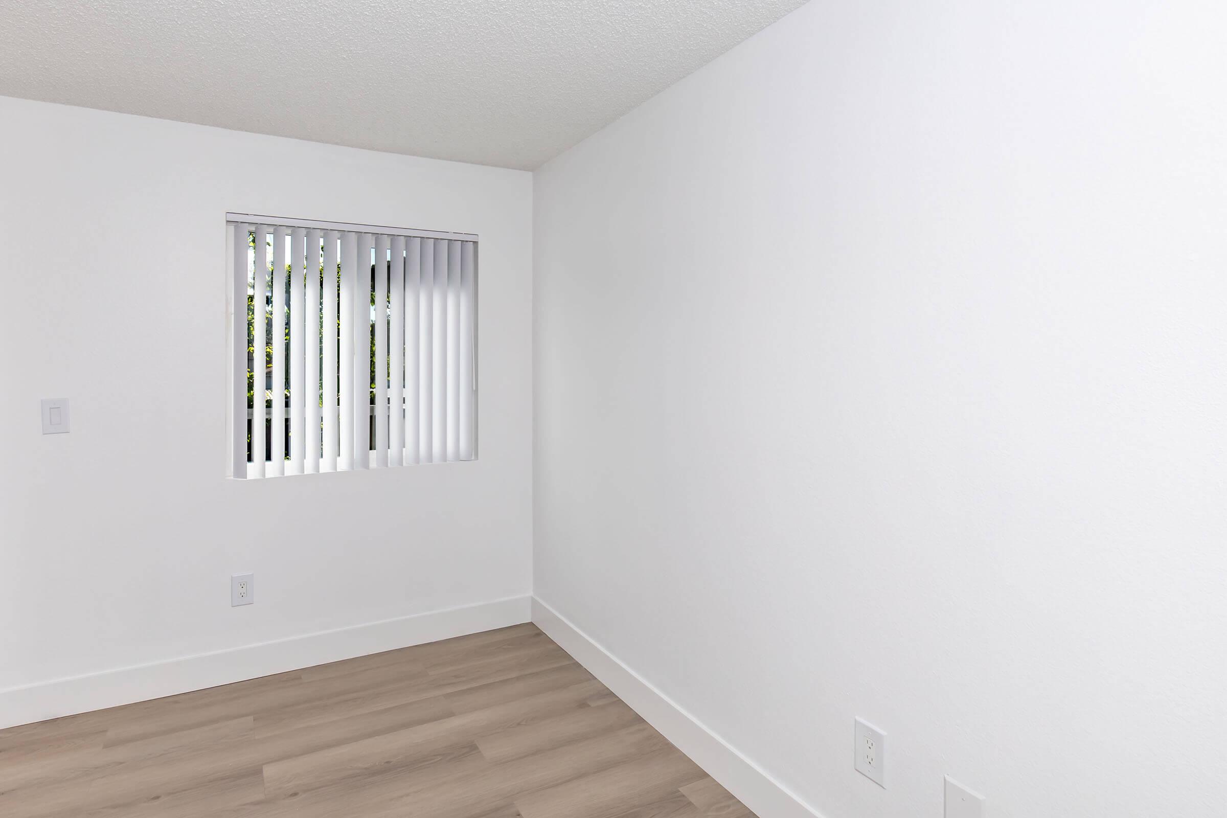 A well-lit, empty room with white walls and light wood flooring. It features a window with vertical blinds, allowing natural light to enter, and minimal furnishings or décor, creating a clean and spacious feel.