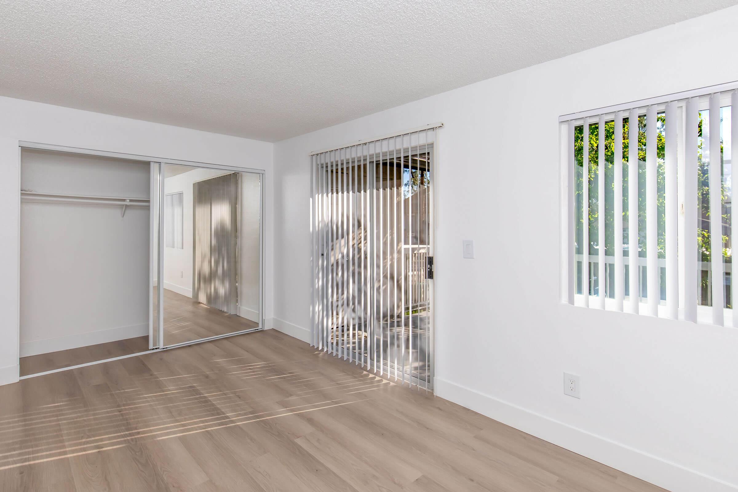 A bright, empty room with light-colored walls and laminate flooring. It features a large sliding door with vertical blinds leading outside, a window with vertical blinds, and a mirrored closet. Soft daylight illuminates the space, creating a welcoming atmosphere.