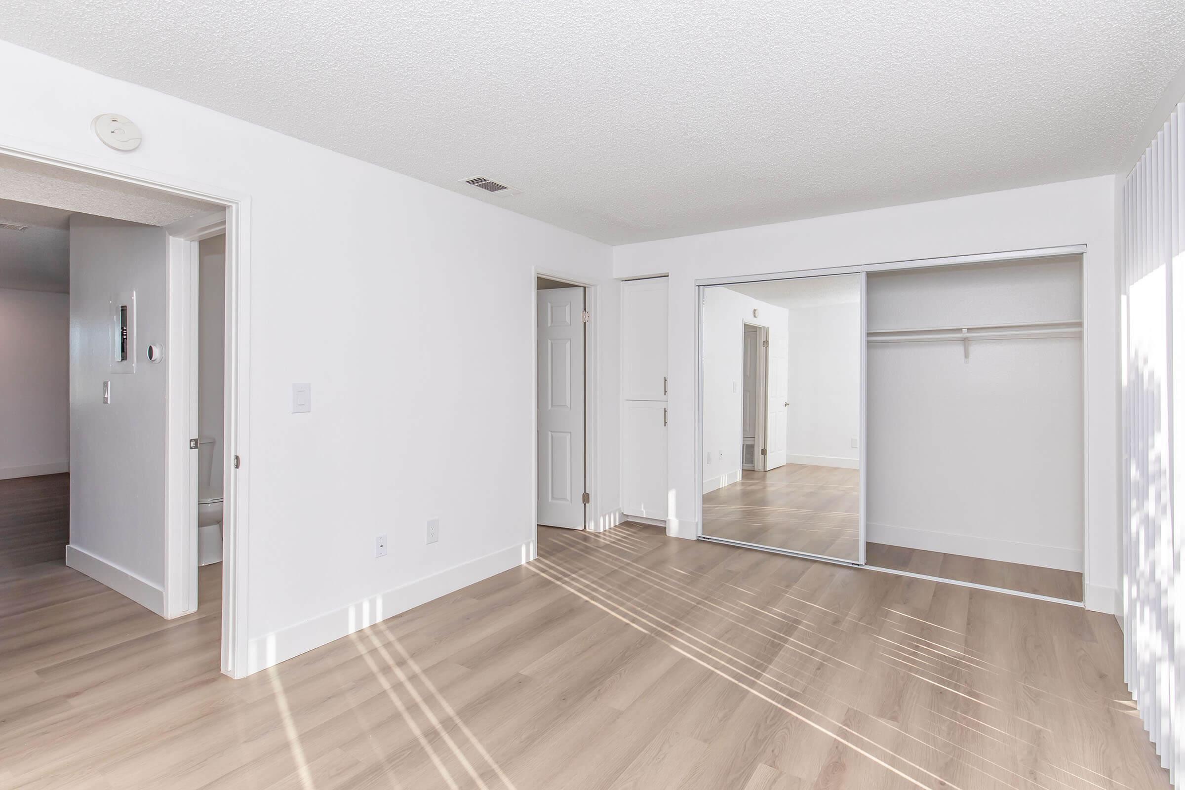 A bright, empty room with light-colored walls and laminate flooring. It features a mirrored closet door and two doors leading to other areas. Sunlight streams in, creating a warm and inviting atmosphere. The room is spacious with a clean, minimalistic design.