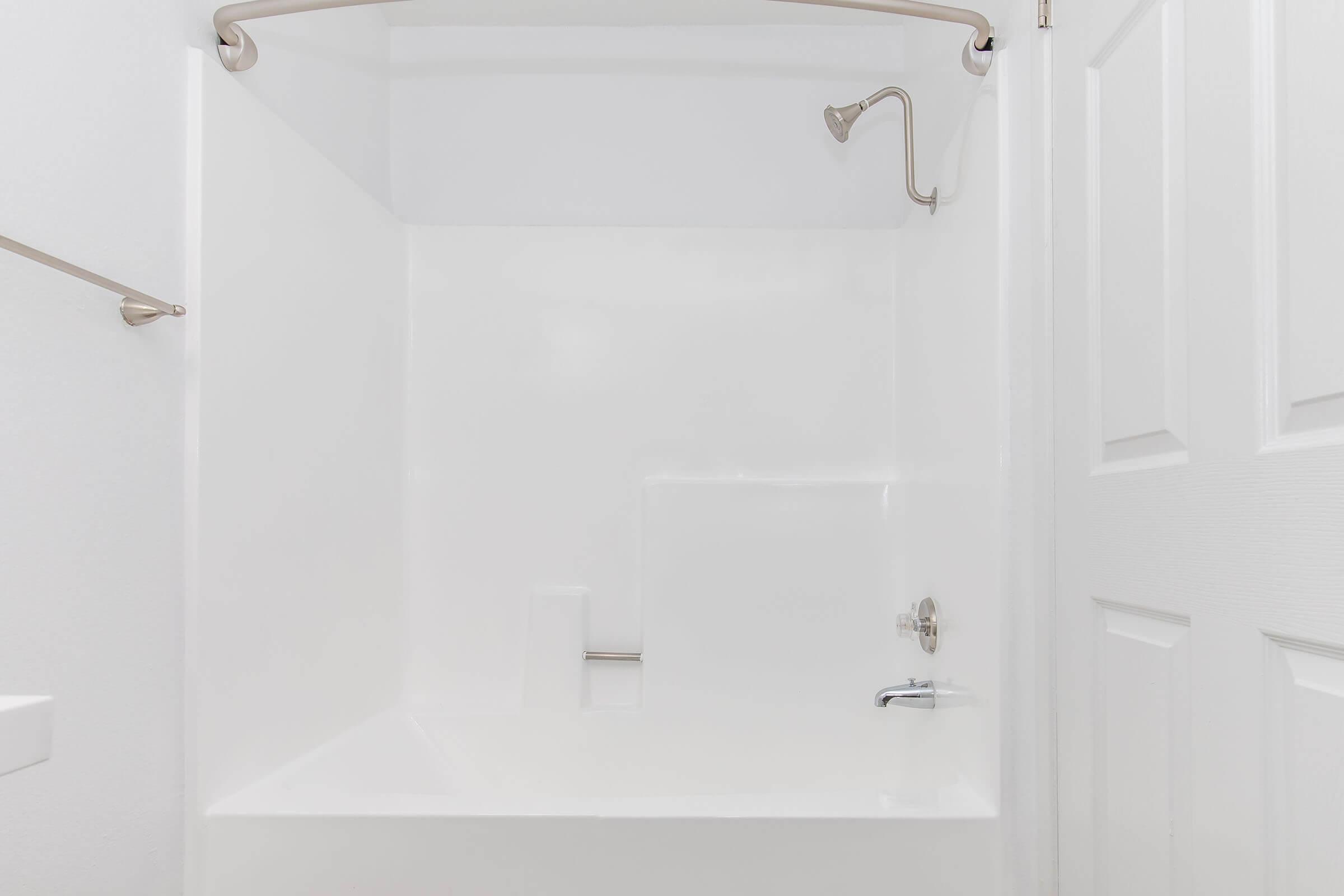 A clean, empty bathroom with a white shower stall, including a showerhead and a faucet. The walls are plain white, and there is a towel bar on one side. A closed door is visible on the right side. The overall look is minimalistic and bright.