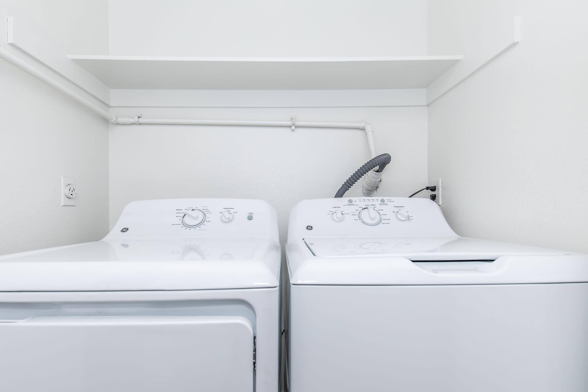 A clean laundry room featuring a white washing machine and a white dryer side by side against a plain wall. There are pipes and a vent connected to the appliances, and a shelf above them. The space is bright and uncluttered, emphasizing a minimalist design.