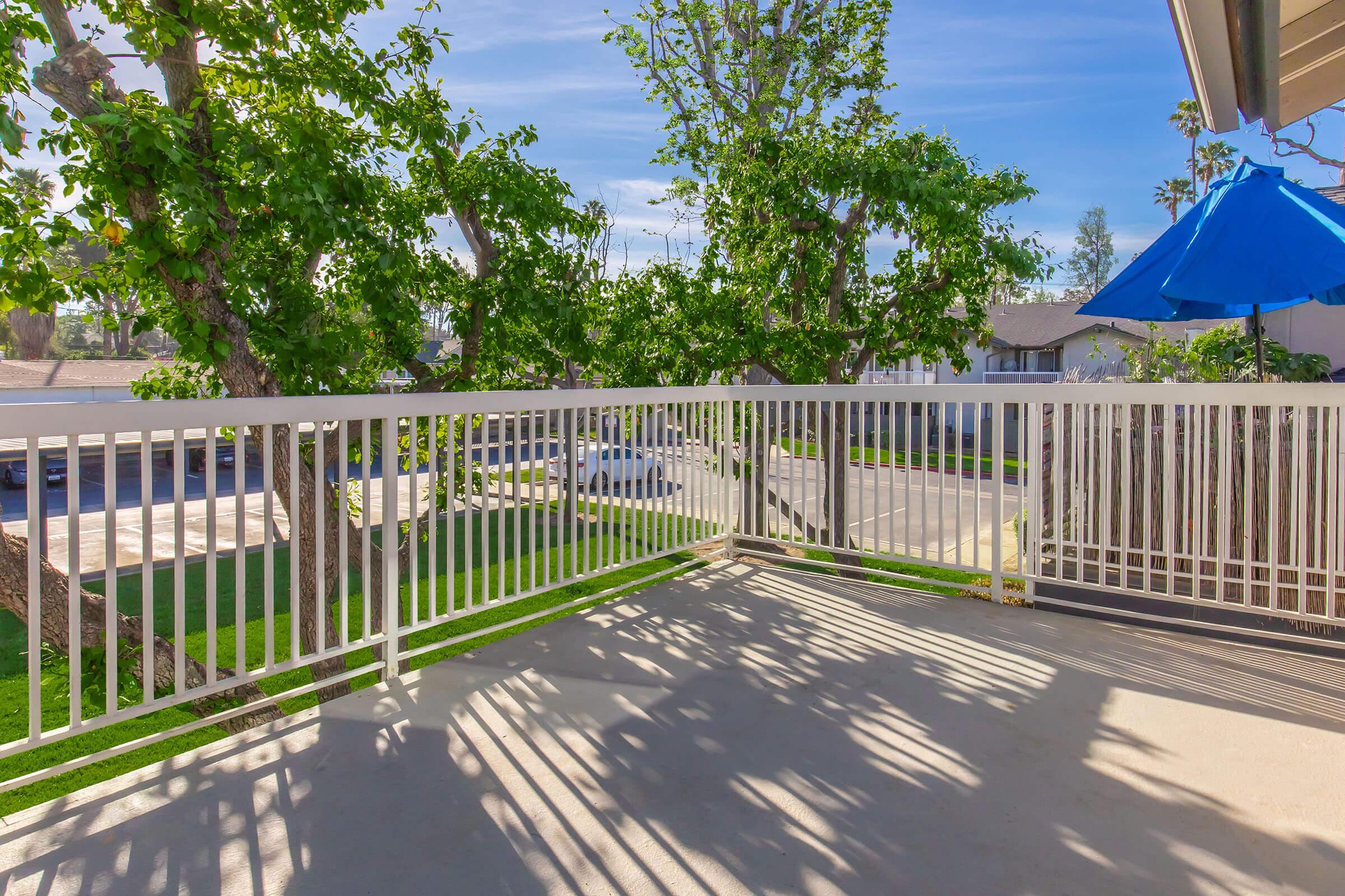 A spacious balcony with a white railing, surrounded by lush green trees. Shadows cast on the floor create a patterned effect. A blue umbrella is visible, providing shade. The scene is bright and inviting, with a view of a parking area and nearby homes.