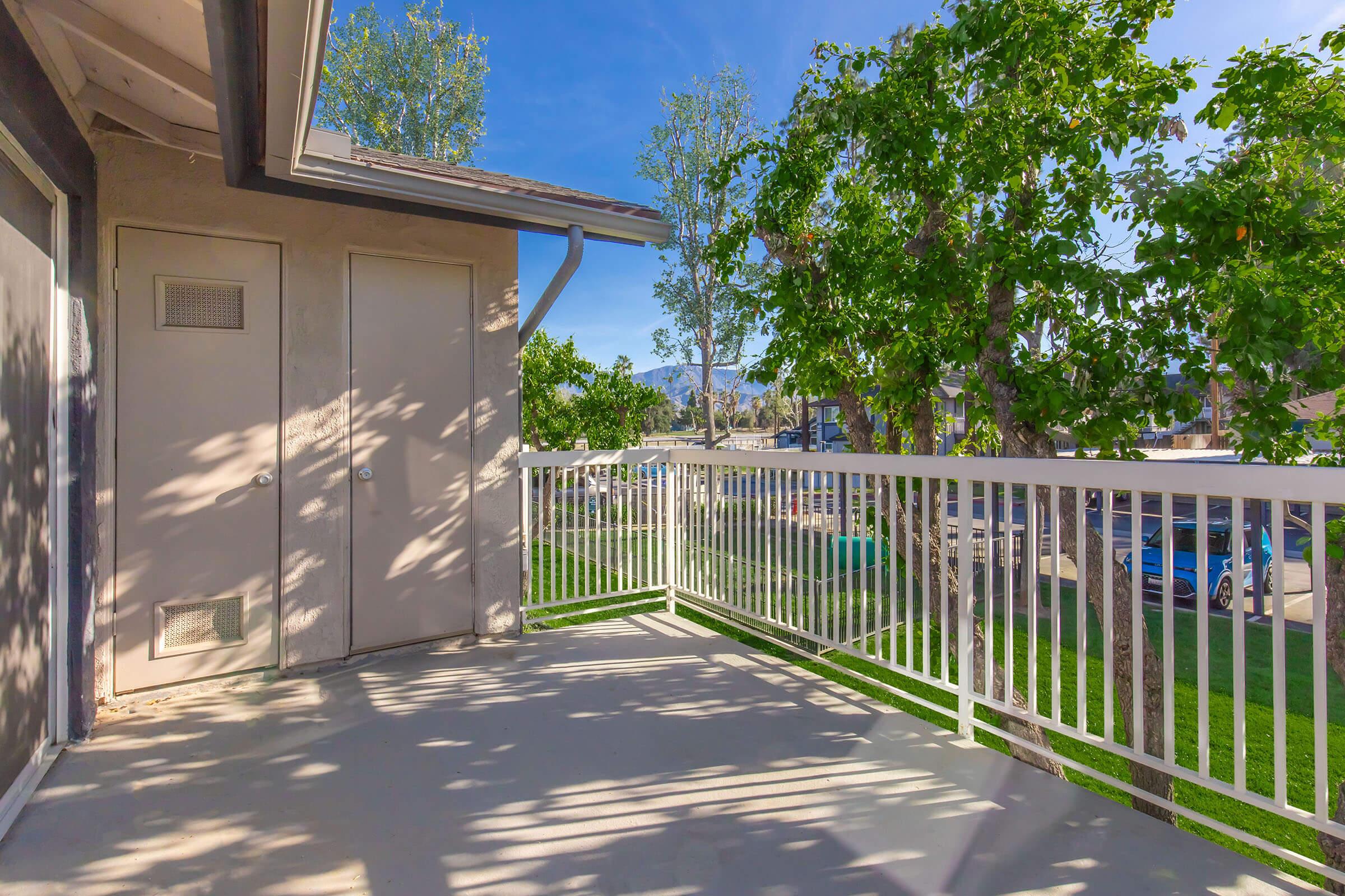 View of a private balcony with a railing, surrounded by green trees and grass. The ground is concrete, and there are two doors on the adjacent wall. Bright blue sky and sunlight create a cheerful and inviting atmosphere. A street with parked cars is visible in the background.
