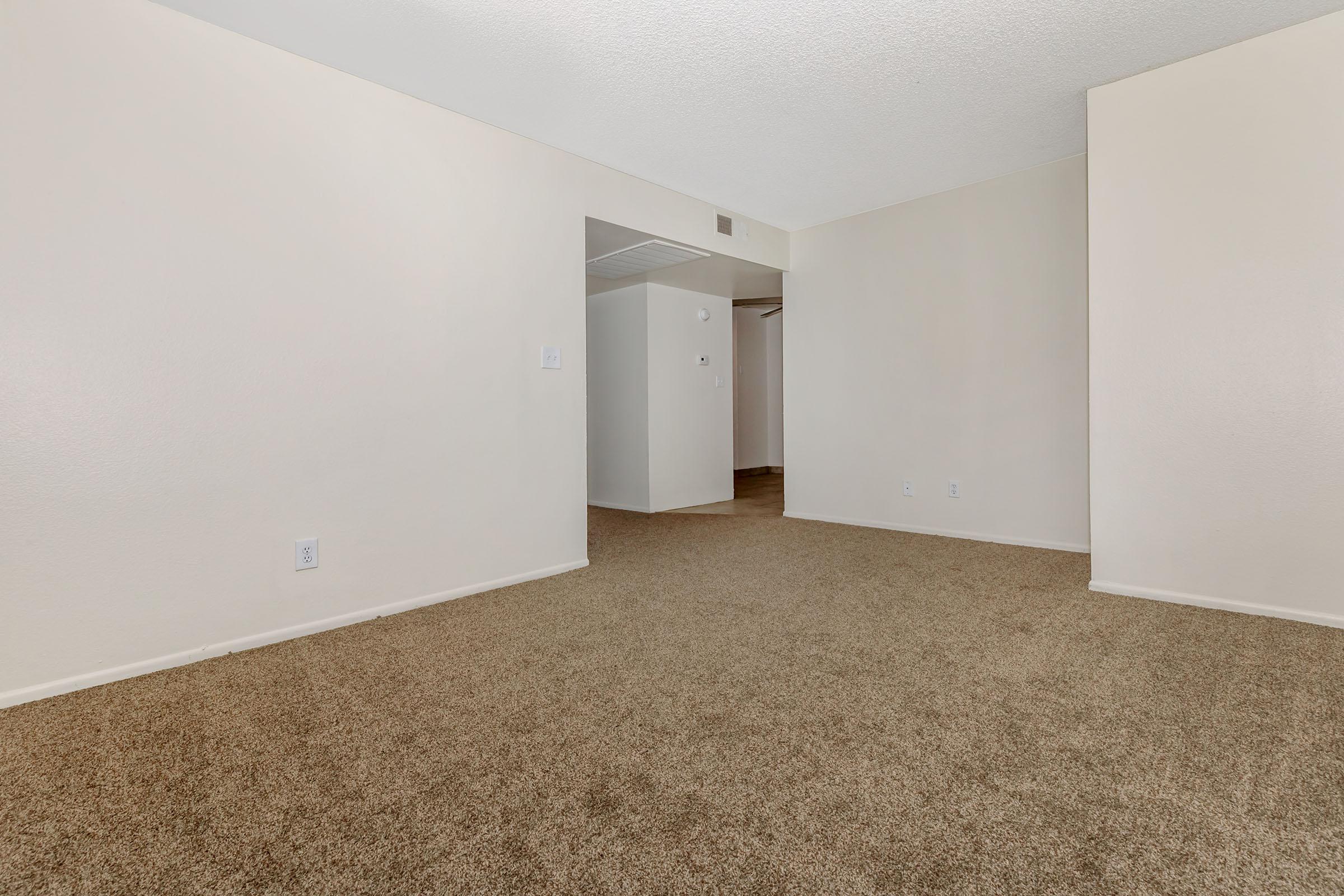 A spacious, empty room with beige walls and brown carpet. There is an open doorway leading to another area, and the lighting is bright, creating a clean and inviting atmosphere. The room is unadorned, ideal for customization or moving in furniture.