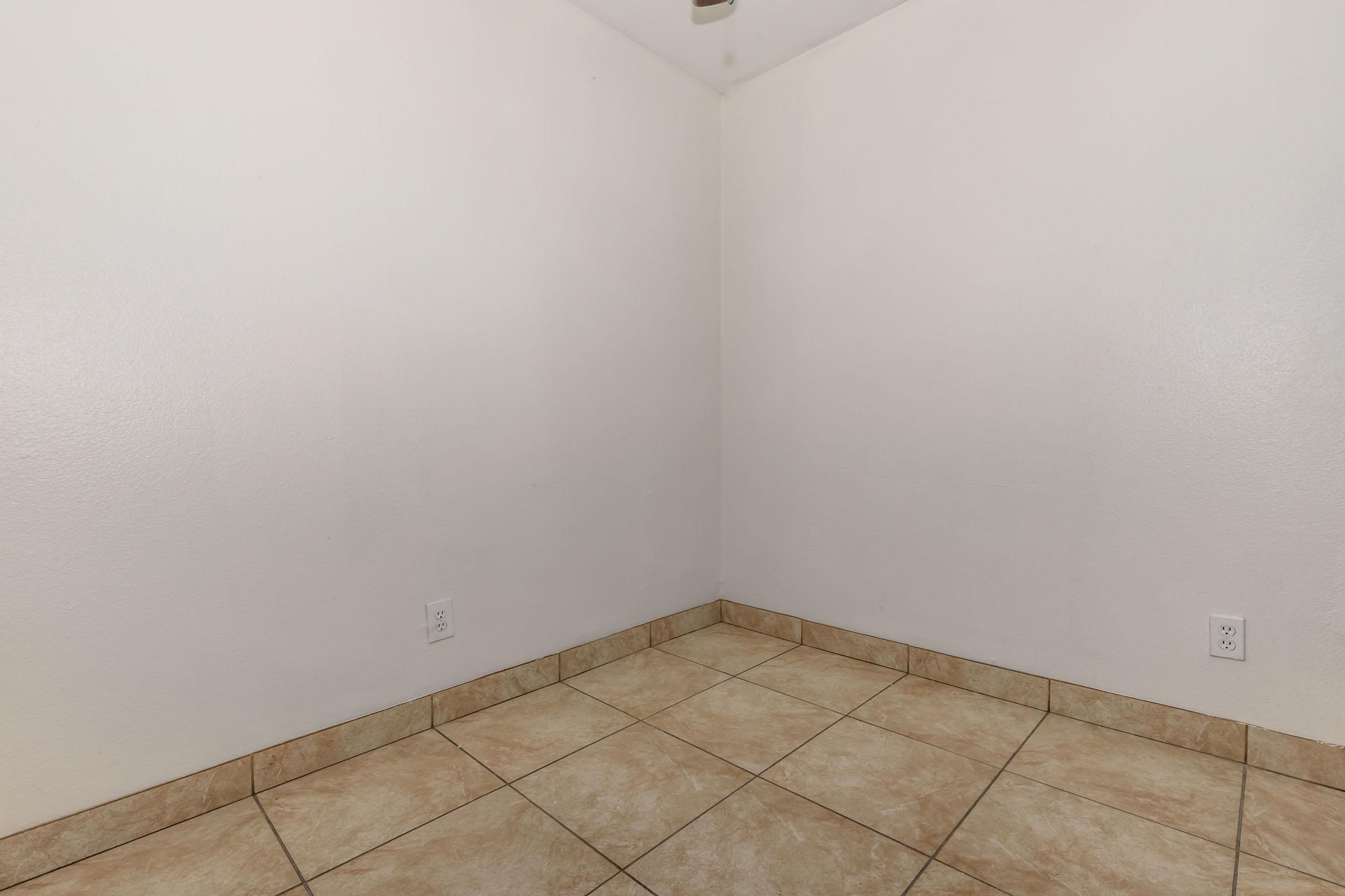 A vacant room featuring white walls and beige tiled flooring. The room has a simple, clean appearance with no furniture or decorations. A ceiling fan is visible, and the space is well-lit, indicating it is ready for use or decoration.