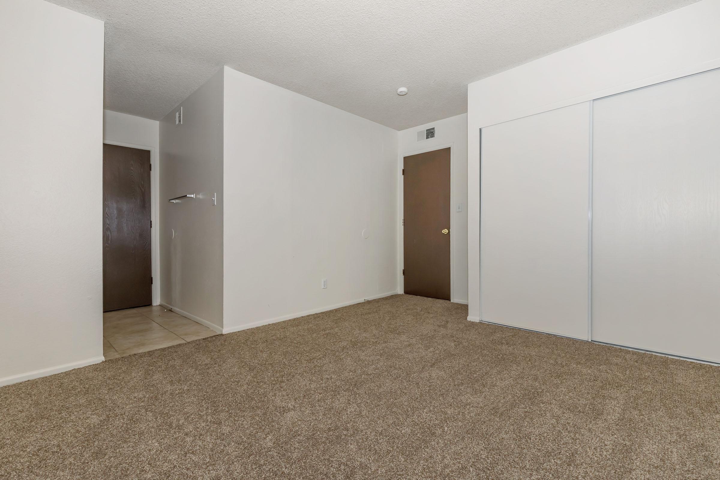 A spacious, empty room with beige carpet and light-colored walls. There is a doorway on the left leading to another room and a closet door on the right. The space is well-lit and features a simple, neutral color palette, suggesting potential for personalization or decoration.