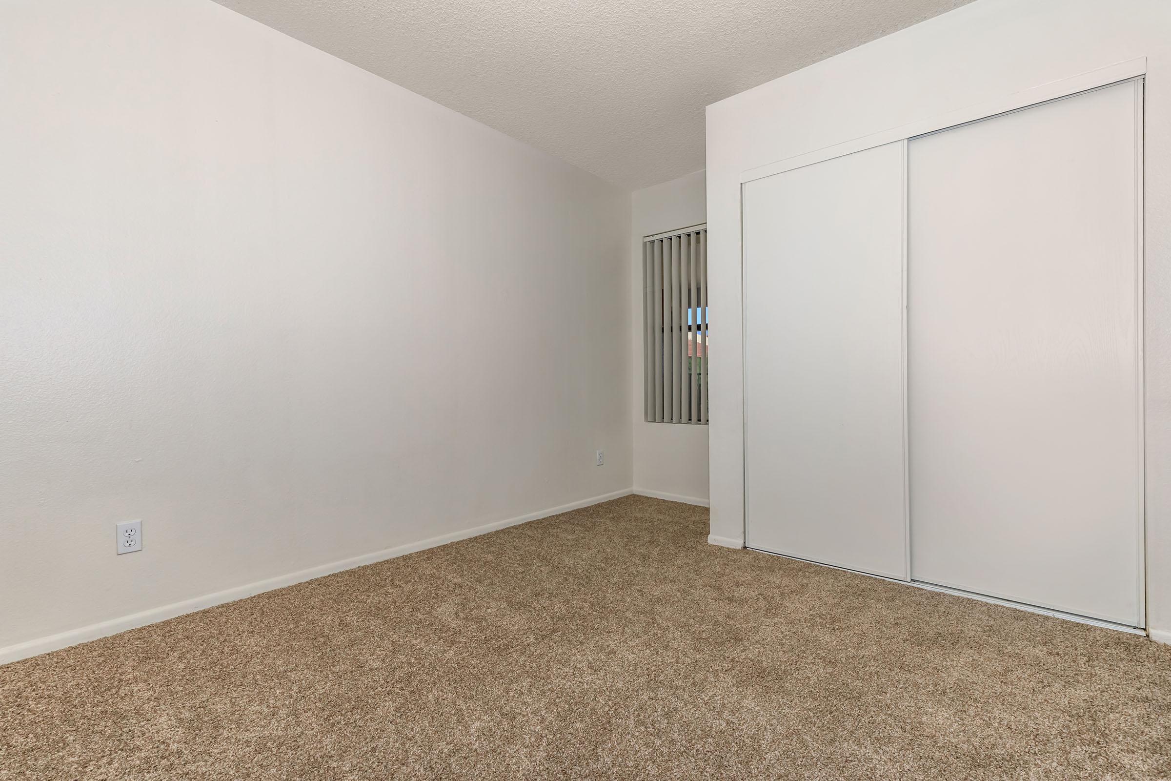 A spacious, empty room with beige carpet flooring and light-colored walls. A white closet with sliding doors is visible on one side. There is a window with vertical blinds allowing natural light to enter. The overall atmosphere is simple and uncluttered, ideal for various uses.