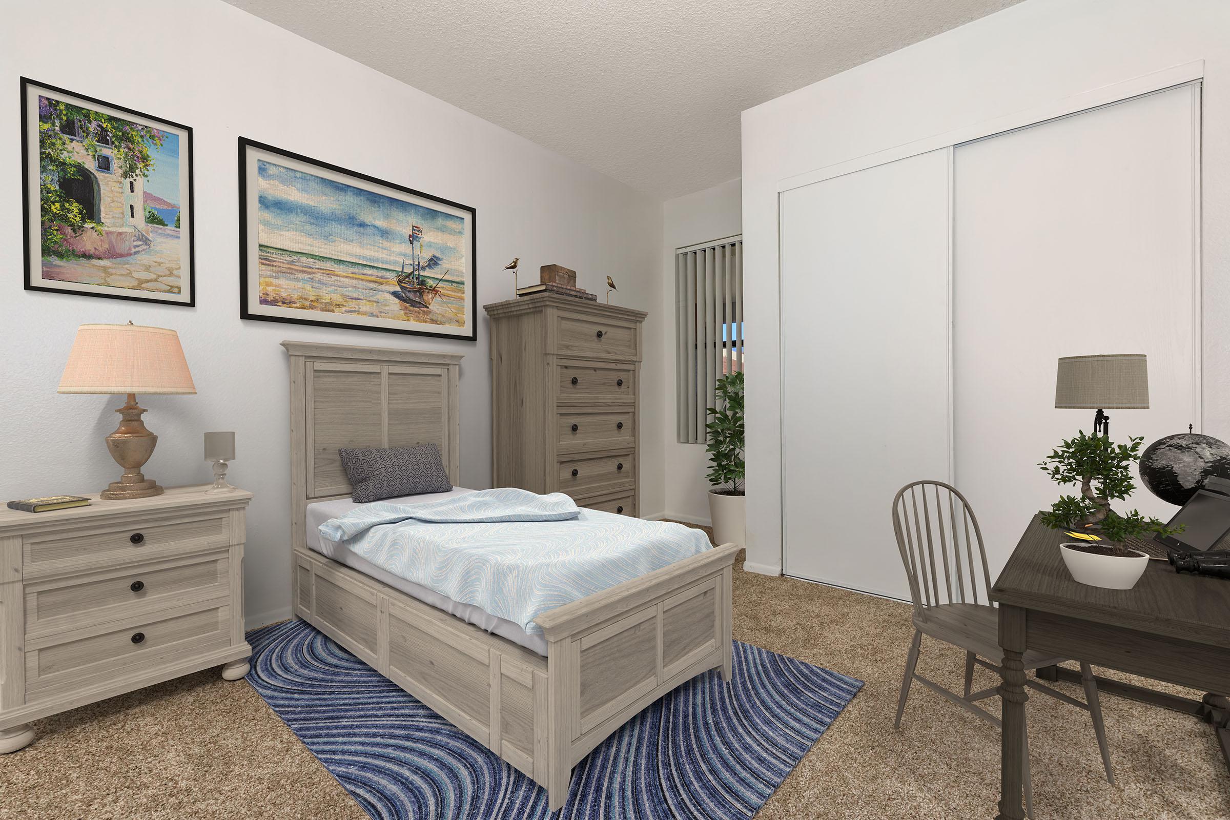 A cozy bedroom featuring a light-colored bed with a blue and white blanket, a wooden nightstand with a lamp, and a tall dresser. The walls are adorned with framed artwork of landscapes. A small desk with a globe is positioned near a plant, and a blue rug adds warmth to the space. A sliding closet door is visible.