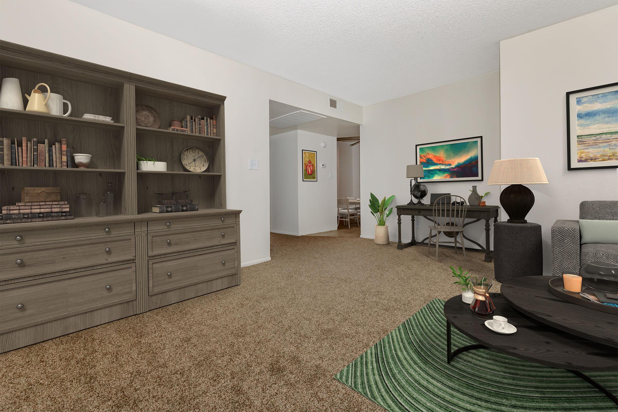 A cozy living space featuring a bookshelf filled with books and decorative items, a coffee table with a plant and drinks, and a comfortable seating area. There’s a desk against the wall and artwork displayed, contributing to a warm, inviting atmosphere. The room has beige walls and brown carpet.