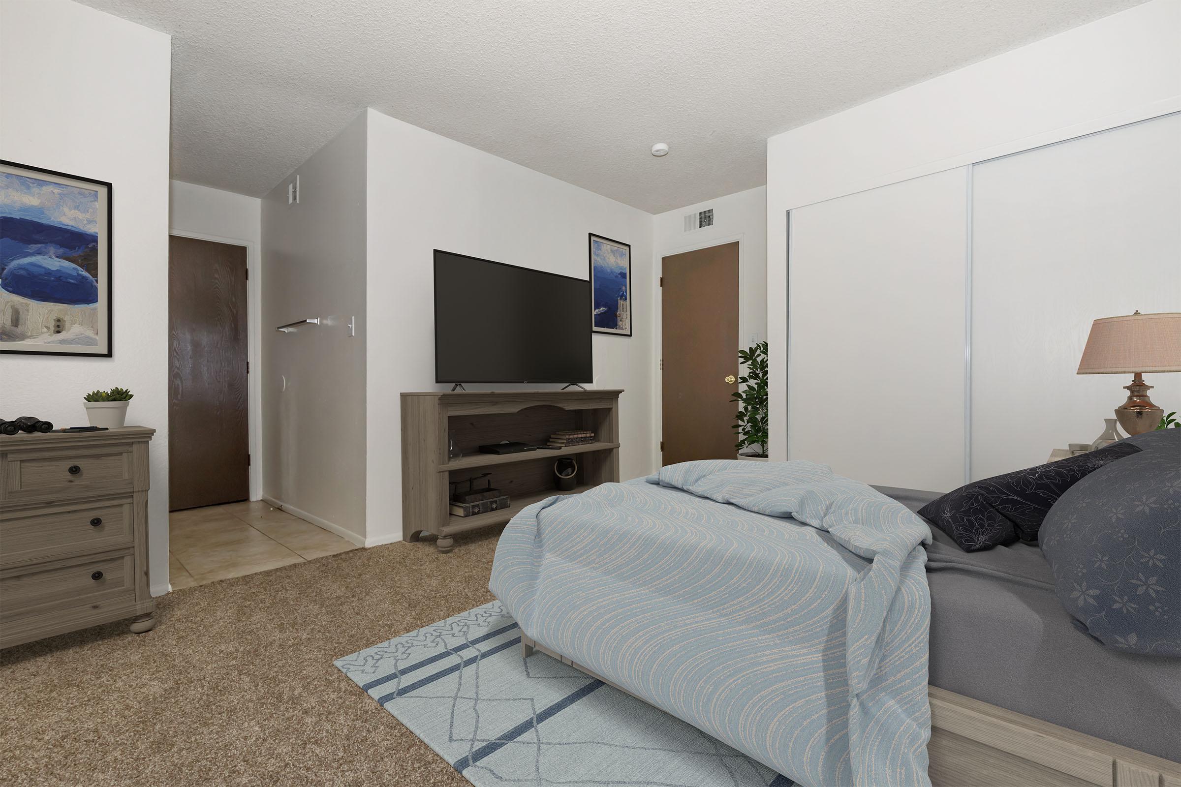 A cozy bedroom with a large bed covered in a light blue duvet. There's a TV on a wooden stand in front of the bed, and framed art on the walls. A door leads to a hallway, and a plant adds a touch of greenery. The carpet is brown, and the lighting is soft, creating a warm atmosphere.