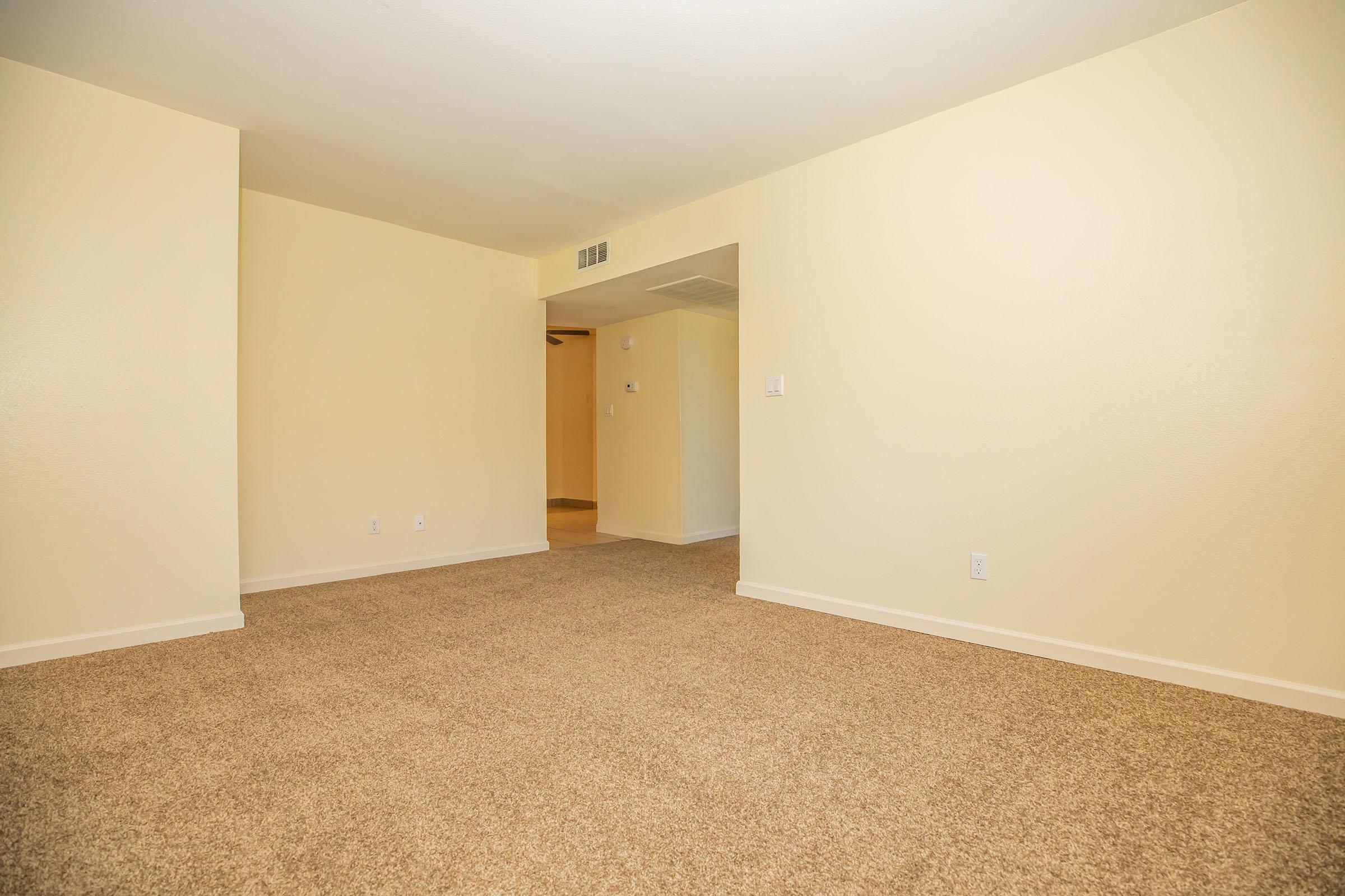 Empty room with light beige walls and tan carpet. The space features an open layout with a doorway leading to another area. Natural light brightens the room, which is free of furniture, emphasizing the spaciousness. The design creates a neutral and inviting atmosphere.