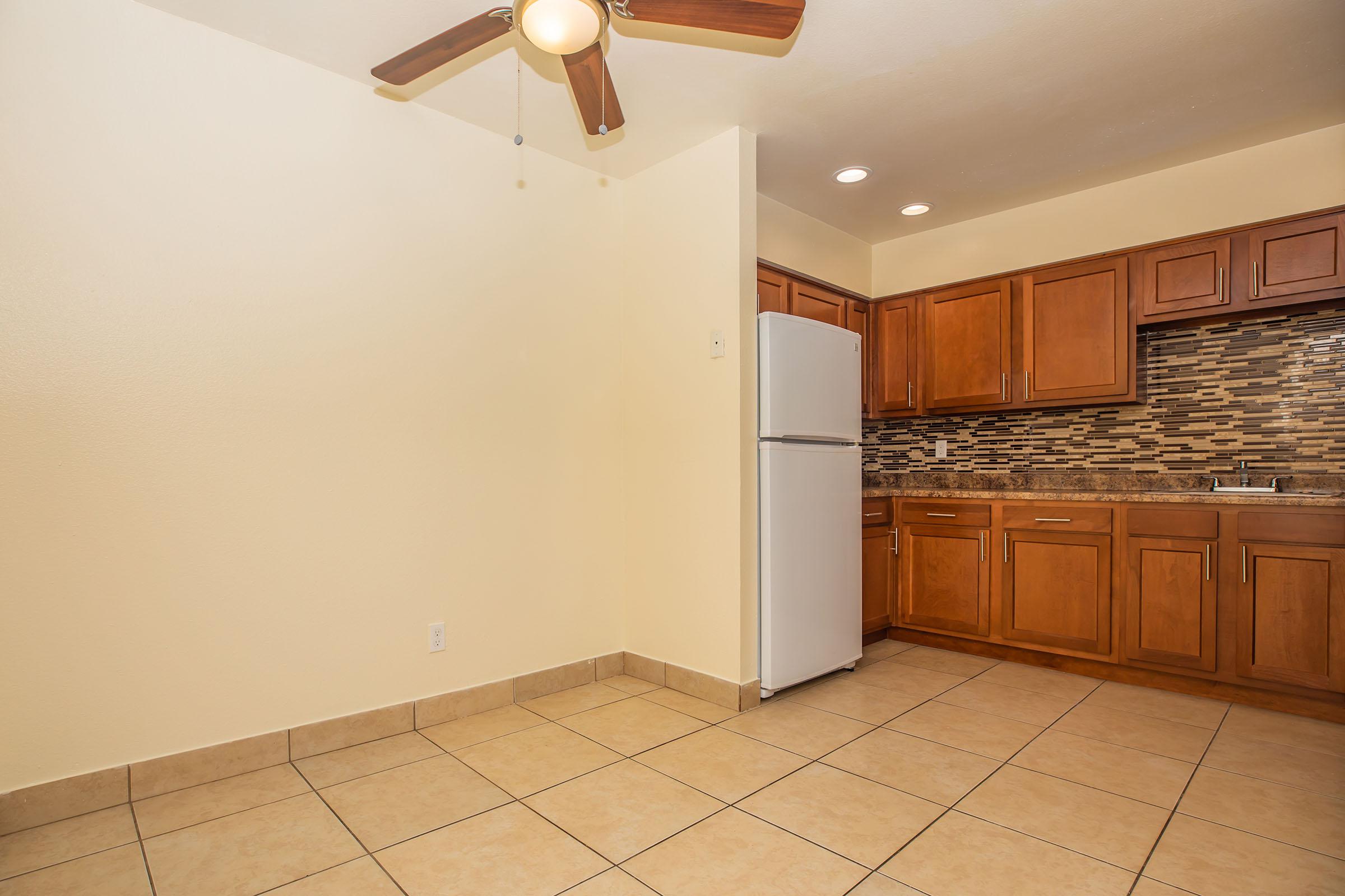 A spacious kitchen featuring wooden cabinets, a white refrigerator, and a decorative backsplash. The room has tiled flooring and a ceiling fan, with soft lighting creating a warm atmosphere. The wall color is light, enhancing the overall brightness of the space.