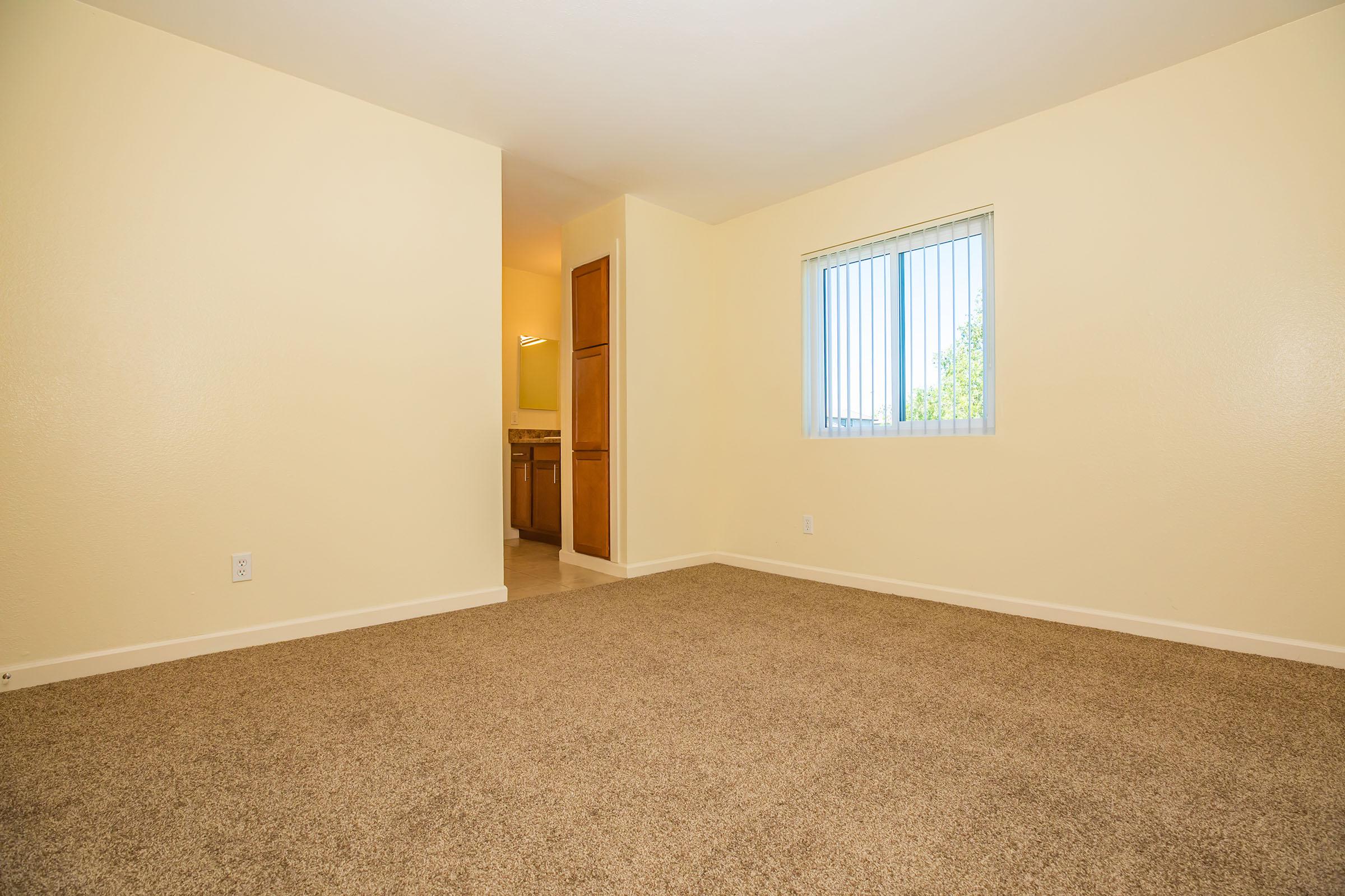 A spacious empty room with light yellow walls and beige carpet. A single window with vertical blinds allows natural light, leading to a small kitchen area partially visible in the background. The room has a minimalistic feel, ideal for decorating or personalizing.