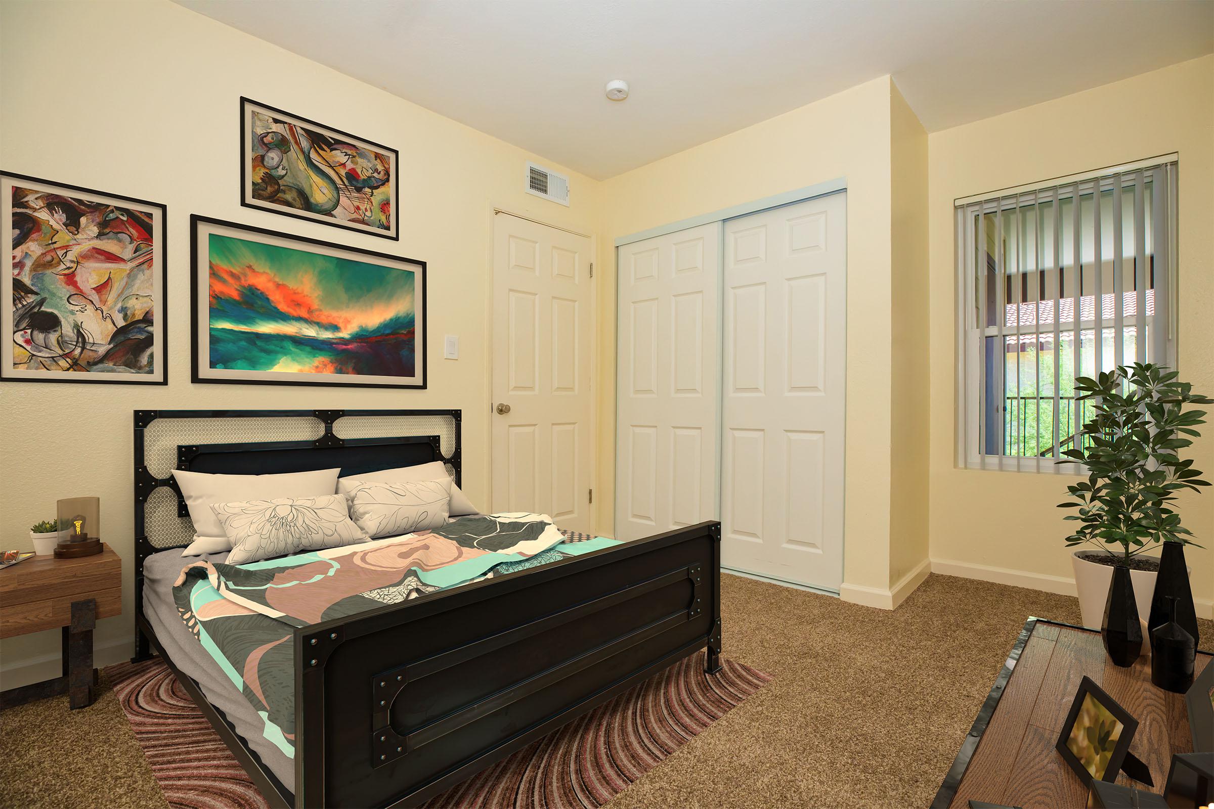 A cozy bedroom featuring a black metal bed frame with colorful bedding, adorned with framed artwork on the walls. There's a side table with a decorative item, a plush rug underfoot, and a large window with blinds allowing natural light to enter. The room has a warm, inviting feel.