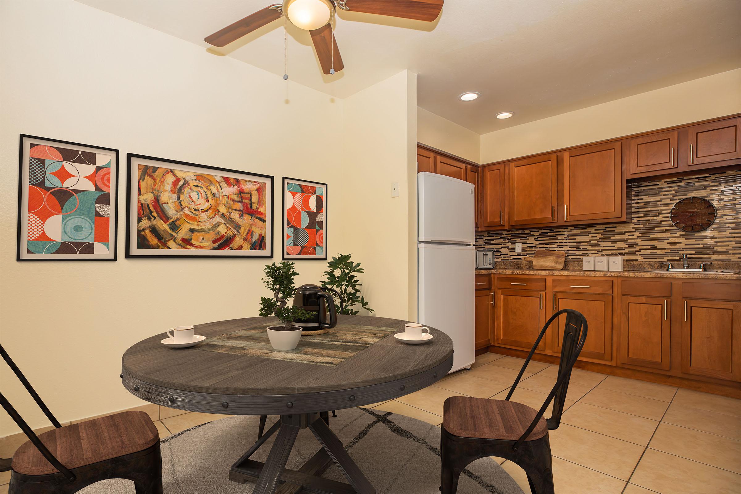 A cozy kitchen area featuring a round wooden dining table with two chairs, a small potted plant, and two coffee cups. The walls are decorated with colorful abstract art. A white refrigerator is positioned beside wooden cabinets, and a tiled backsplash enhances the modern aesthetic of the space.