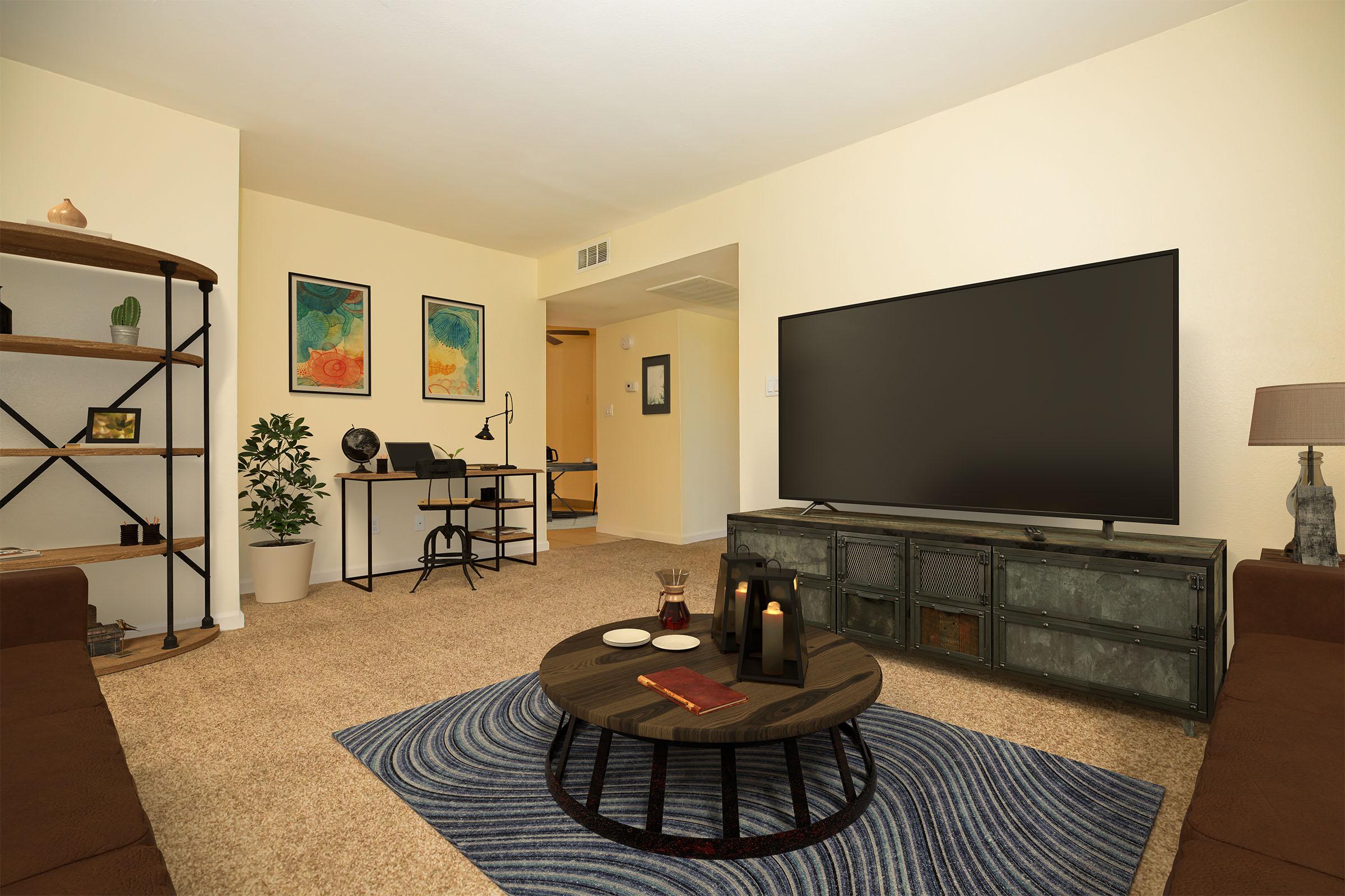 A cozy living room featuring a large flat-screen TV on a dark console, a round coffee table with candles and books, and a patterned rug. There is a bookshelf with decorative items, a small plant, and a desk with a chair. Two framed artworks hang on the wall, and the space is brightly lit with neutral-colored walls and carpet.