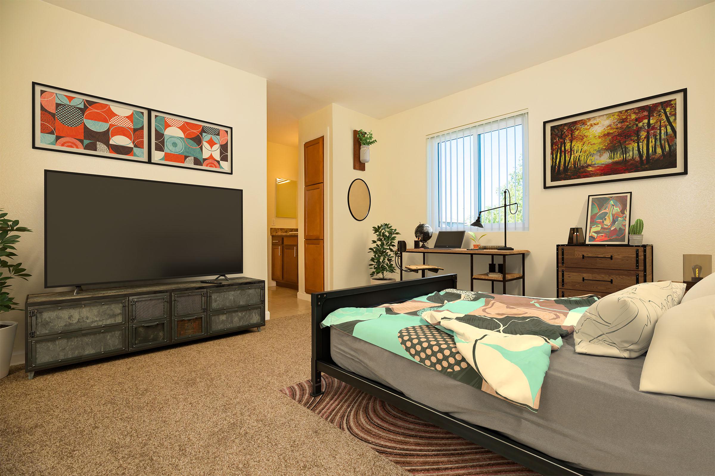 A cozy, modern bedroom featuring a bed with a colorful quilt, a large television on a stylish cabinet, a desk by the window with a chair, and decorative plants. Walls are adorned with framed artwork, and there's a wooden dresser in the corner, creating a warm and inviting atmosphere.