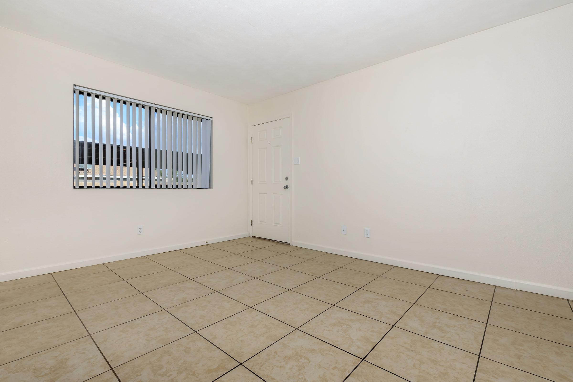 Empty room with beige tile flooring and light-colored walls. A window with vertical blinds allows natural light to enter. The door is visible on one side, and there is no furniture or decoration in the space.