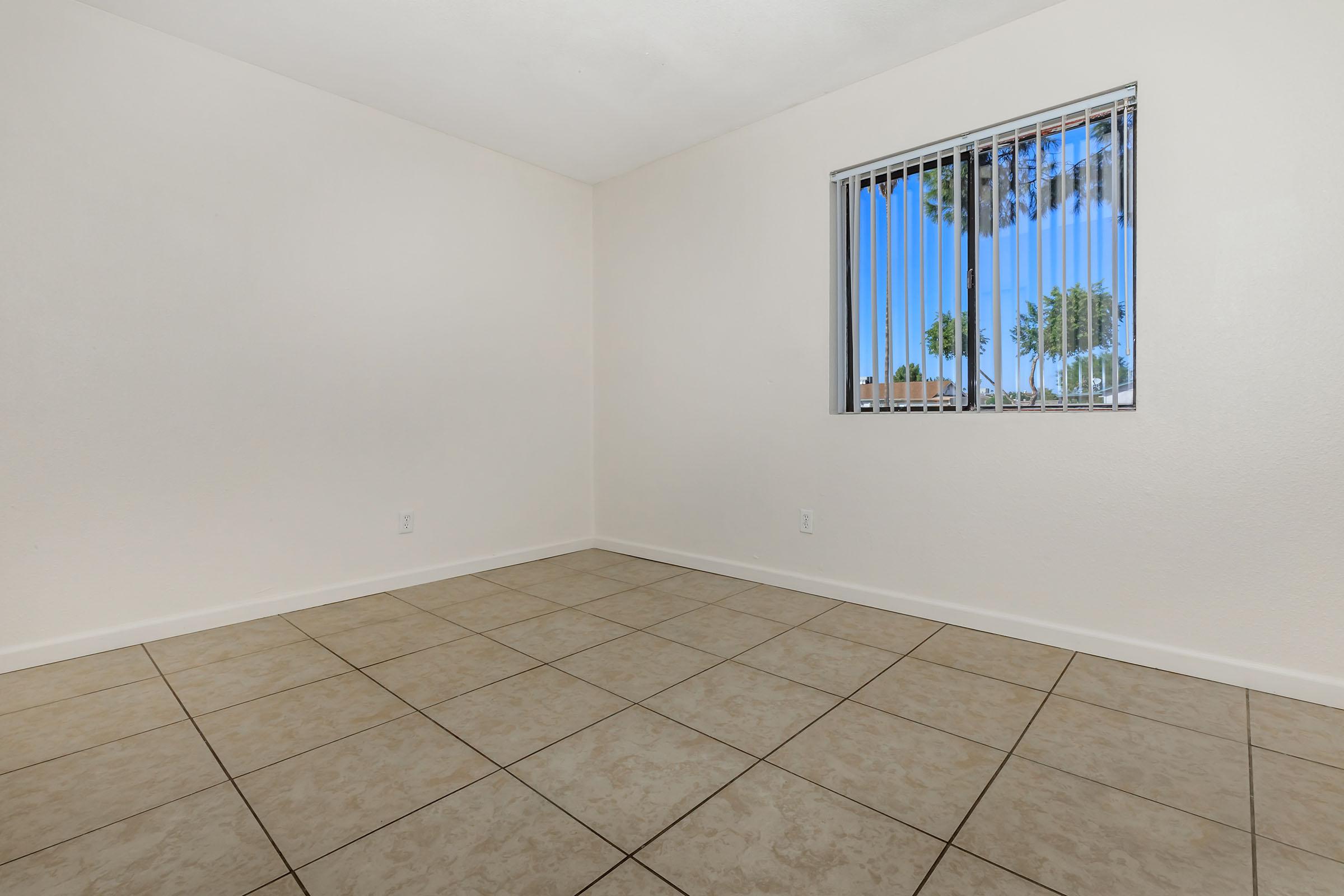 A spacious, empty room with beige tile flooring and white walls. There is a window on the left side featuring vertical blinds, allowing natural light and a view of palm trees outside. The ceiling is plain, enhancing the room's simplicity and openness.