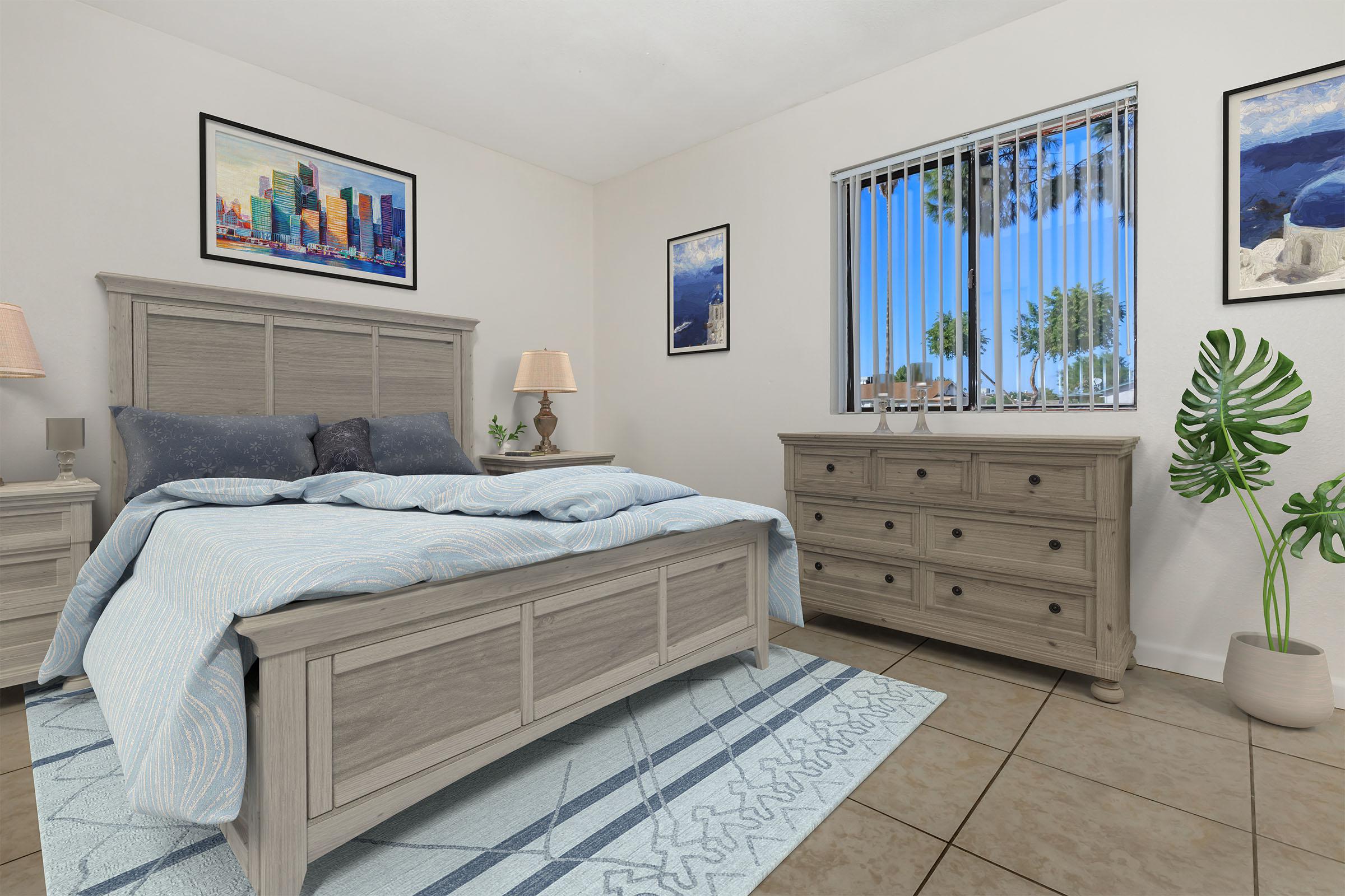 A cozy bedroom featuring a light wooden bed with blue and white bedding, matching nightstands with lamps, and a dresser. The walls are adorned with framed pictures, and a large window lets in natural light, revealing palm trees outside. A decorative plant adds greenery to the space, enhancing the inviting atmosphere.