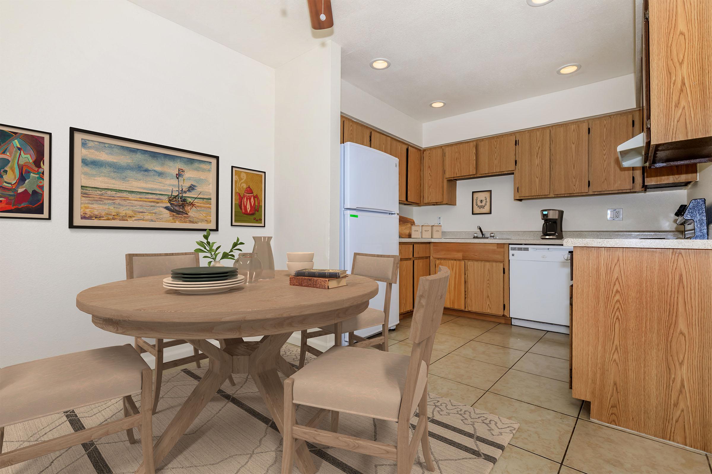 A cozy kitchen featuring a round wooden table with four chairs, a white refrigerator, and wooden cabinetry. The walls are adorned with colorful artwork, and there are appliances like a dishwasher and coffee maker visible. The floor is tiled, and the room is well-lit, creating a warm atmosphere.