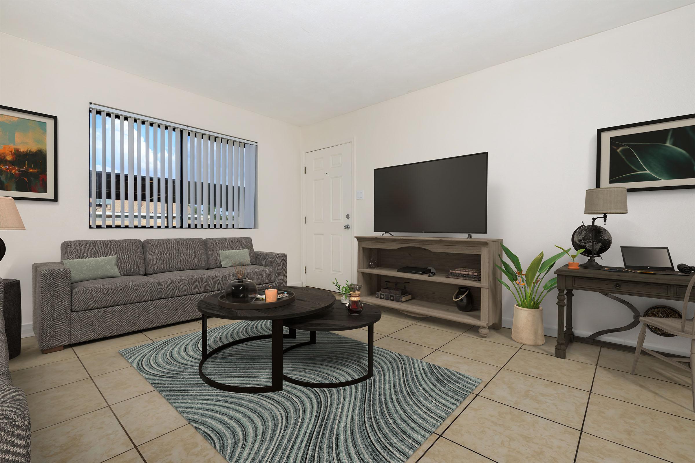 A cozy living room featuring a gray sofa, circular coffee table, and a large TV on a wooden stand. The space includes decorative items like a plant and a globe, with two framed artworks on the walls and natural light coming through the window with vertical blinds. The floor is tiled in neutral colors.