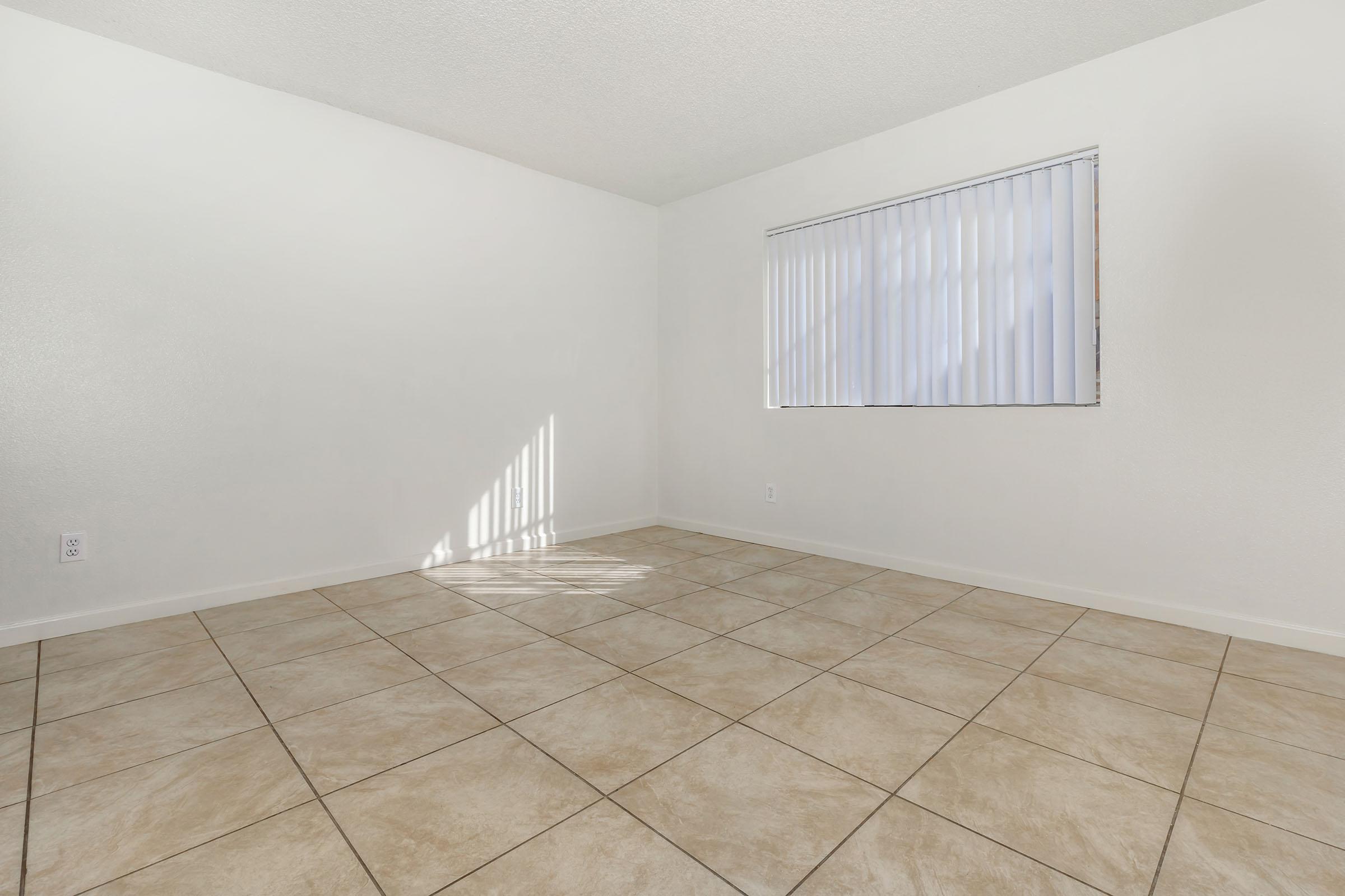 A clean, empty room with beige tiled flooring and white walls. There's a window with vertical blinds allowing natural light to enter, casting a shadow on the floor. The space is bright and spacious, ideal for potential furnishings or decoration.